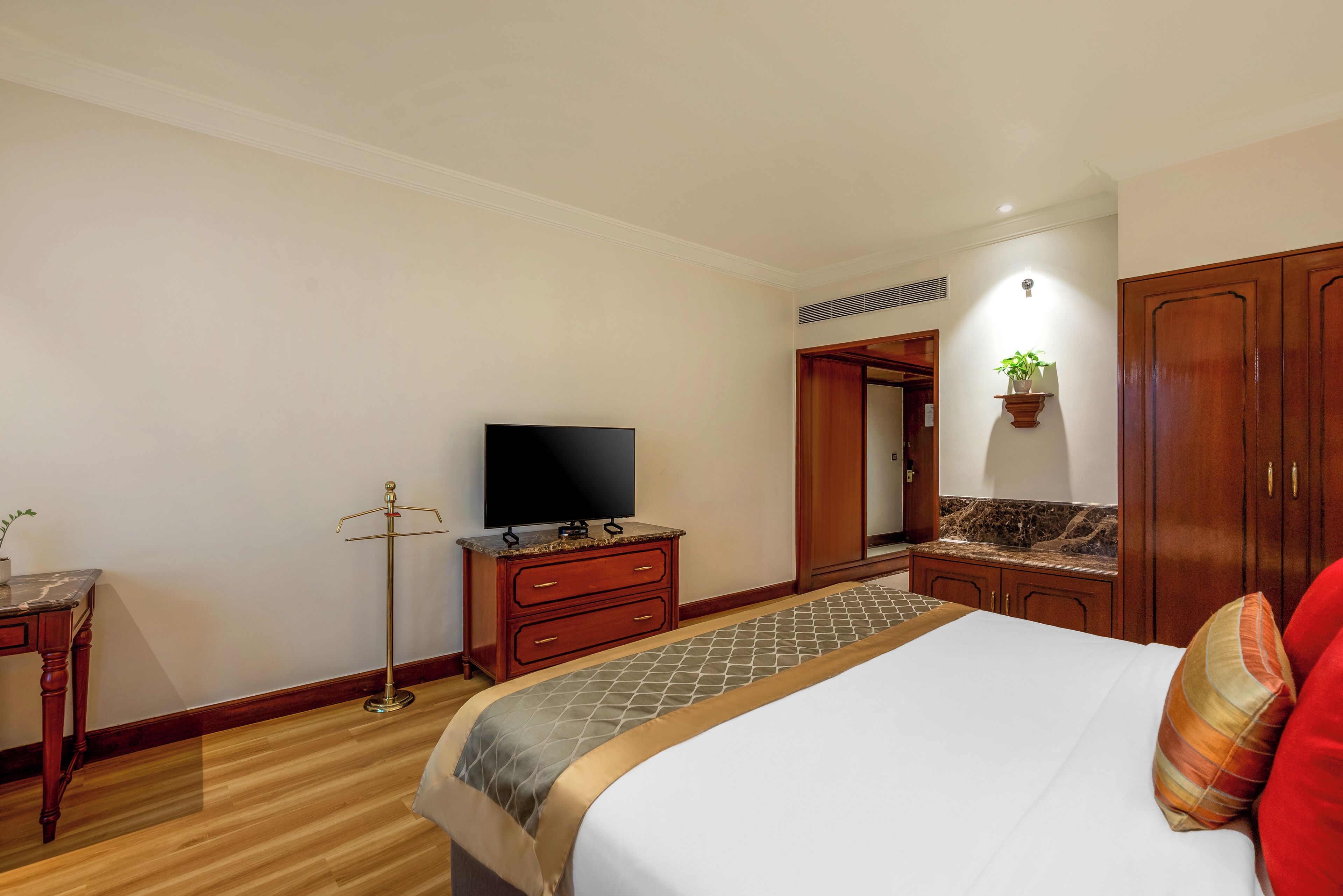 A hotel room with a bed, a television on a wooden dresser, a small table with a plant, and a mirror on the wall.