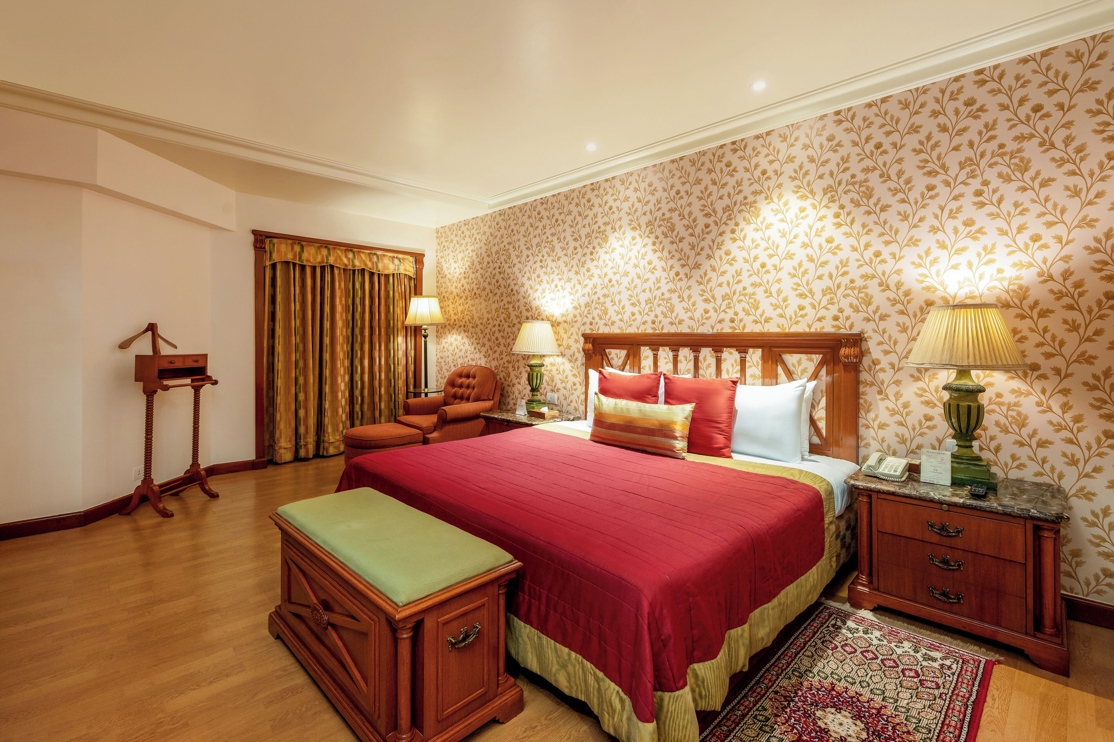 A hotel room with a large bed, wooden nightstands, a green bench, and a patterned wallpaper.