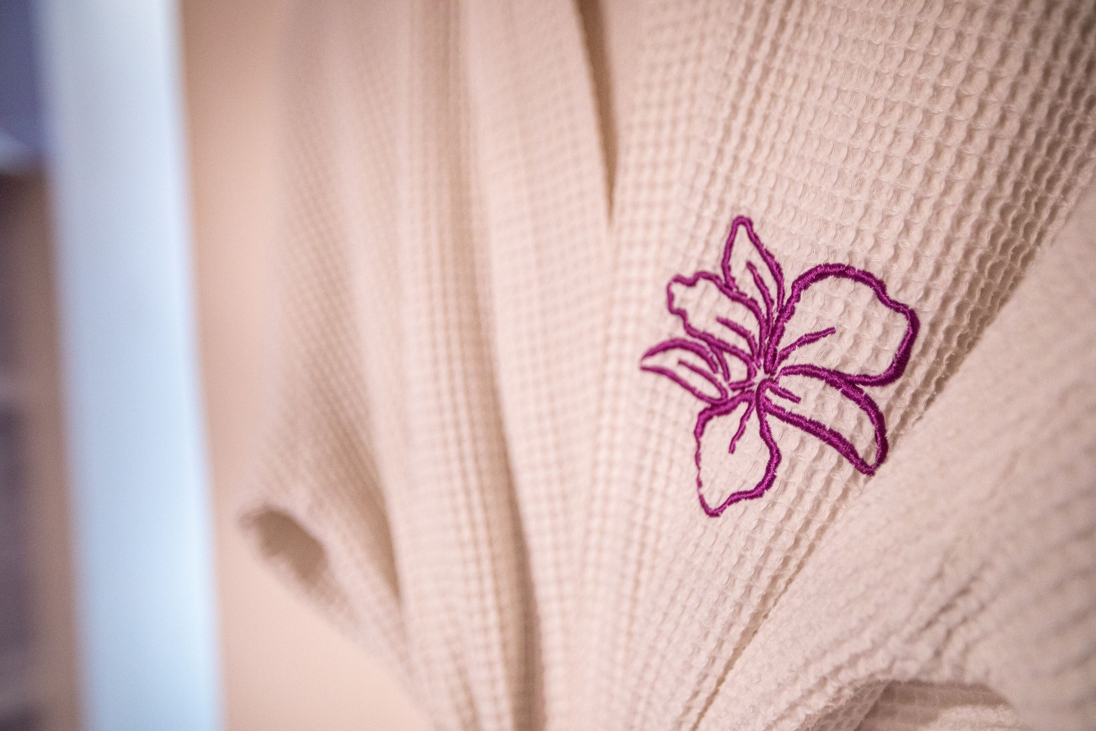 Ein Detailaufnahme eines beige farbenen, strukturierten Stoffes mit einer violett bestickten Blume.