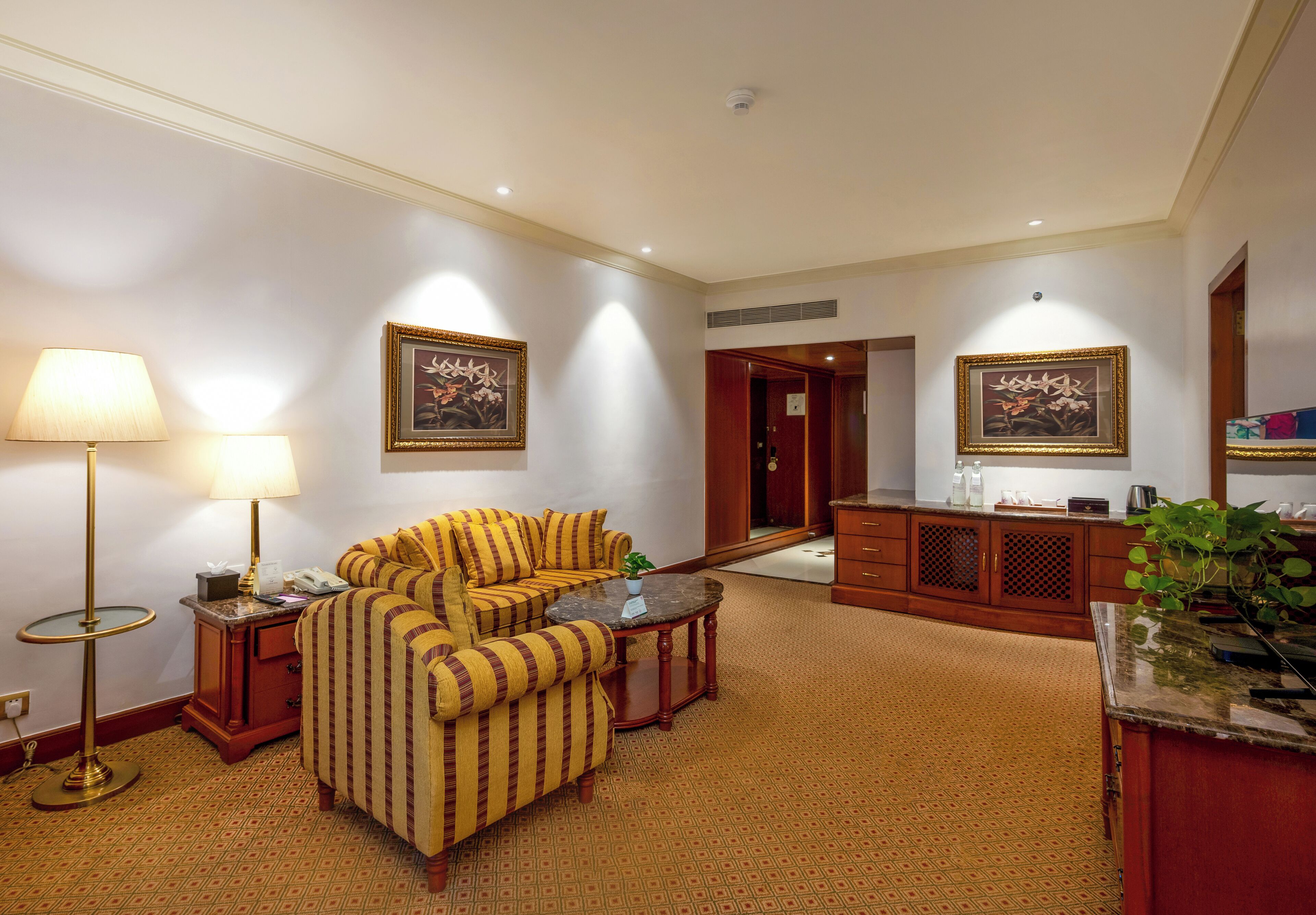 A hotel room with a striped sofa, a small round table, a lamp, a cabinet, and two framed pictures on the walls.