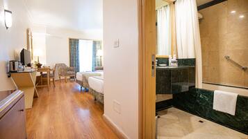 A hotel room with a wooden floor, a bed, a desk with a chair, a television, and a bathroom with a shower and a marble countertop.