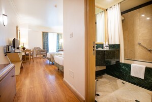 A hotel room with a wooden floor, a bed, a desk with a chair, a television, and a bathroom with a shower and a marble countertop.