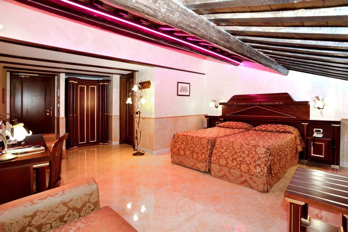 A hotel room with a bed, a desk, and a chair. There is a wooden door and a mirror on the wall.