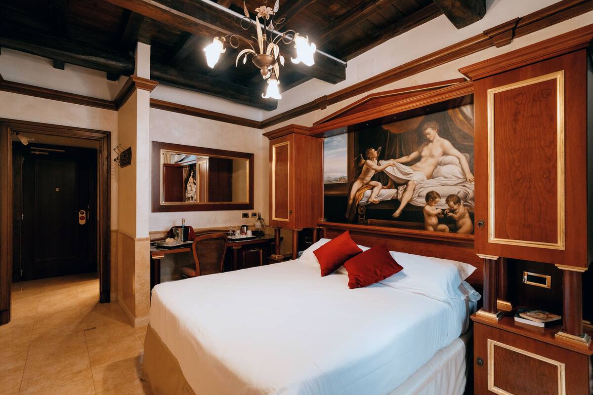 A hotel room with a large bed, wooden paneling, a chandelier, a desk with a chair, and a painting on the wall.