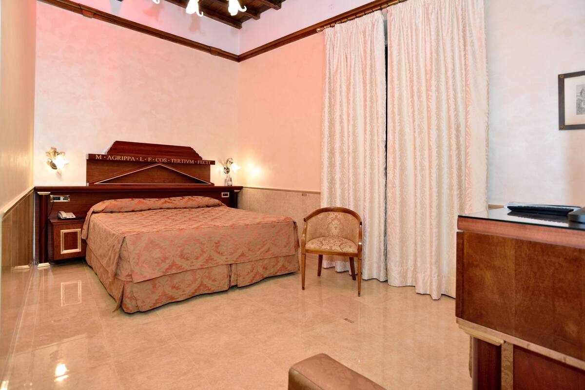 A hotel room with a large bed, a wooden nightstand with a phone, a chair, a desk, and a large window with curtains.