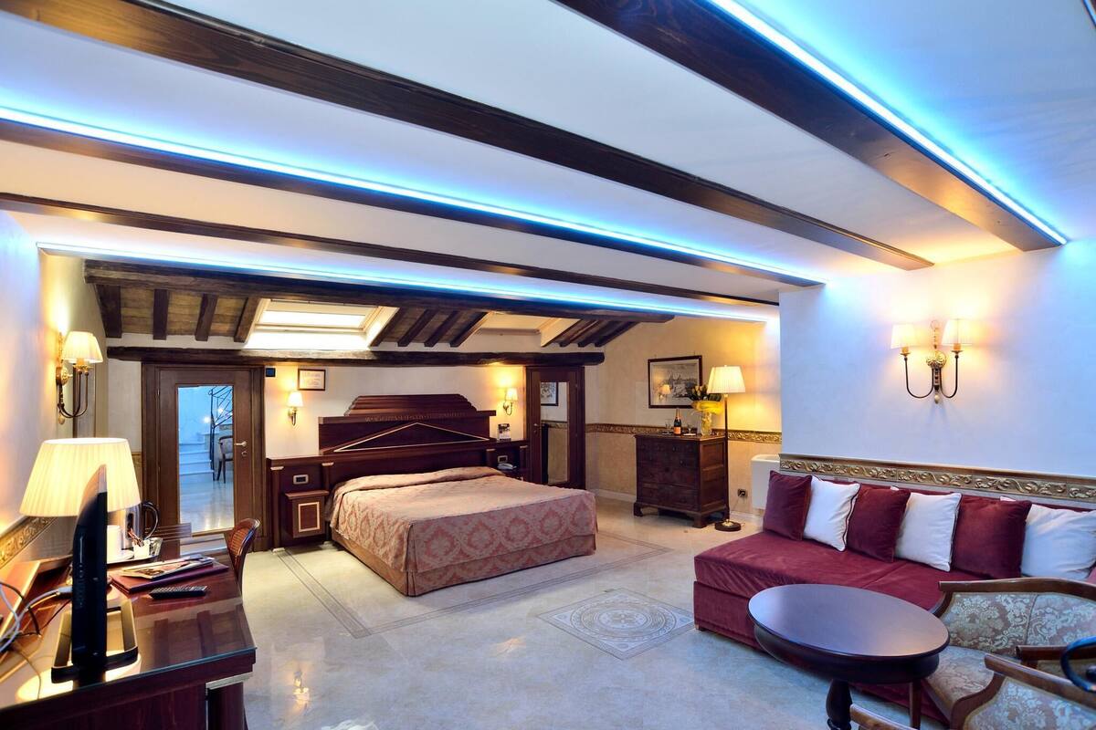 A hotel room with a bed, a sofa, a desk, and a TV. There is a door leading to a balcony.