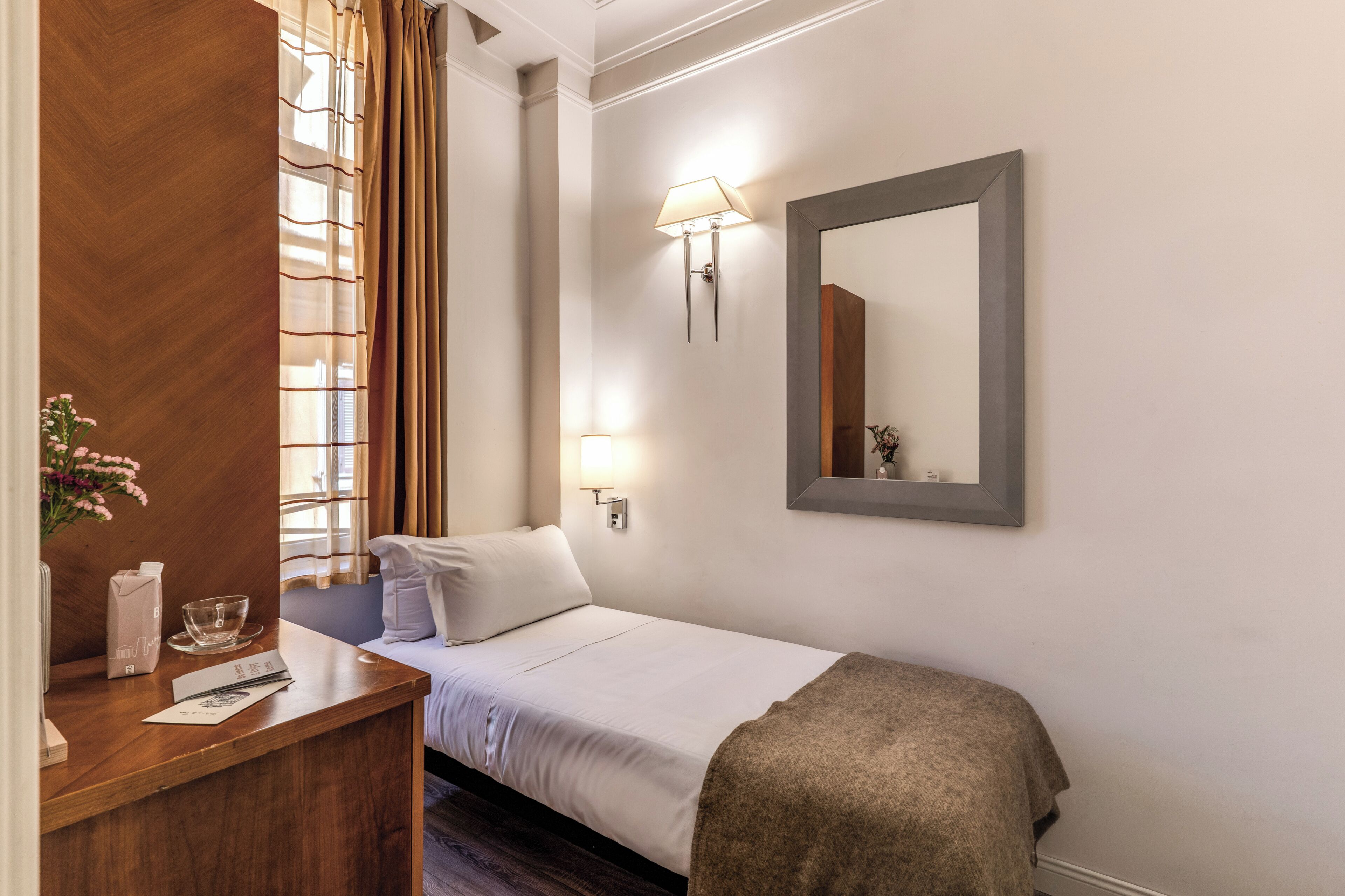 A hotel room with a bed, bedside lamps, a wooden desk, a mirror, and a window with curtains.