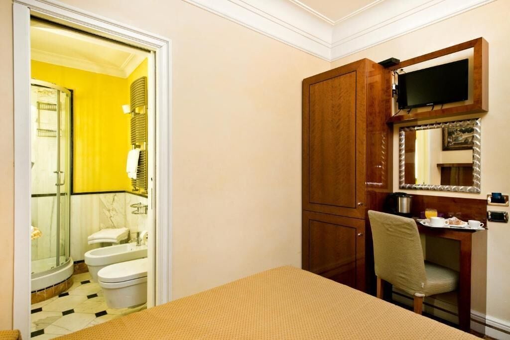 A hotel room with a bed, a desk with a chair, a television mounted on the wall, and a bathroom with a shower and toilet.