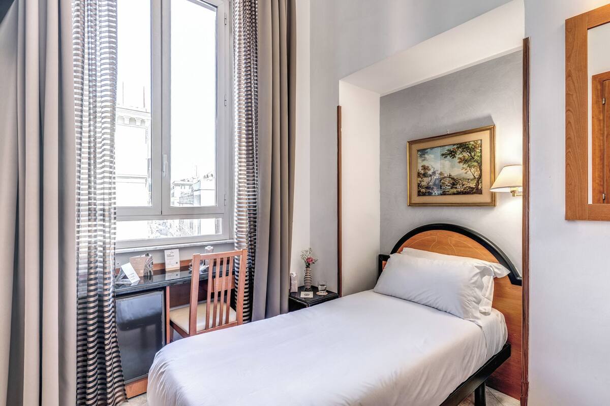 A hotel room with a bed, a desk, a chair, a nightstand, a lamp, and a painting on the wall.