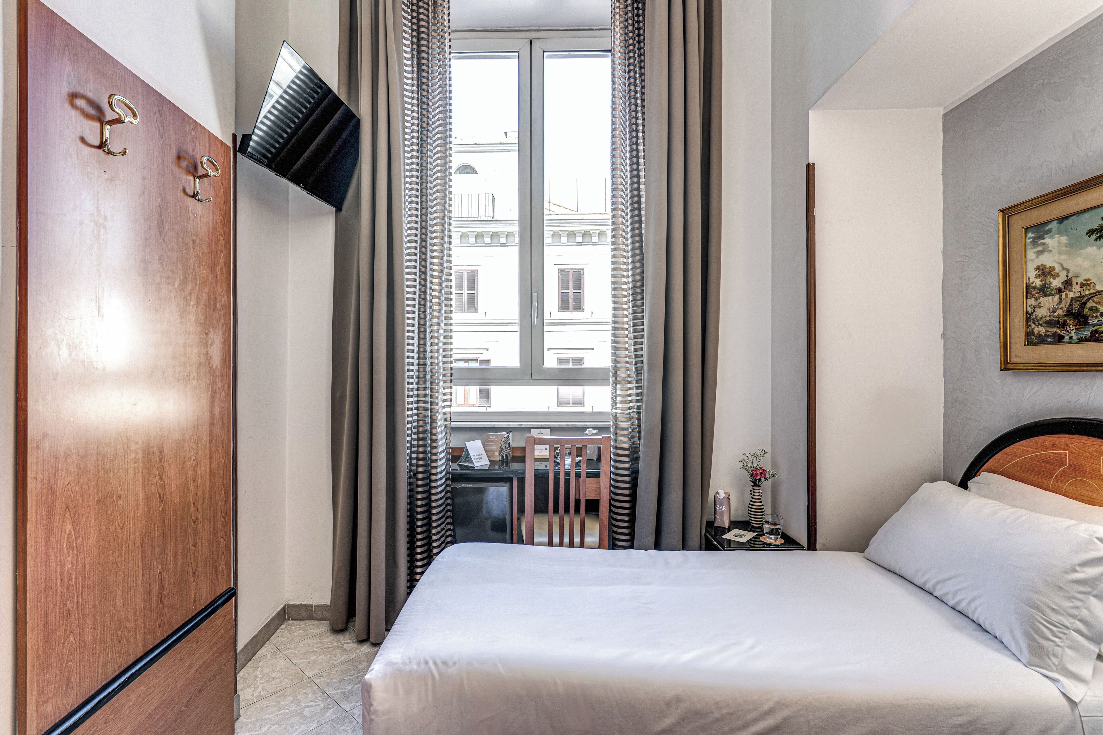 Habitación de hotel con cama, armario de madera, escritorio con silla, televisor montado en la pared y ventana con cortinas.