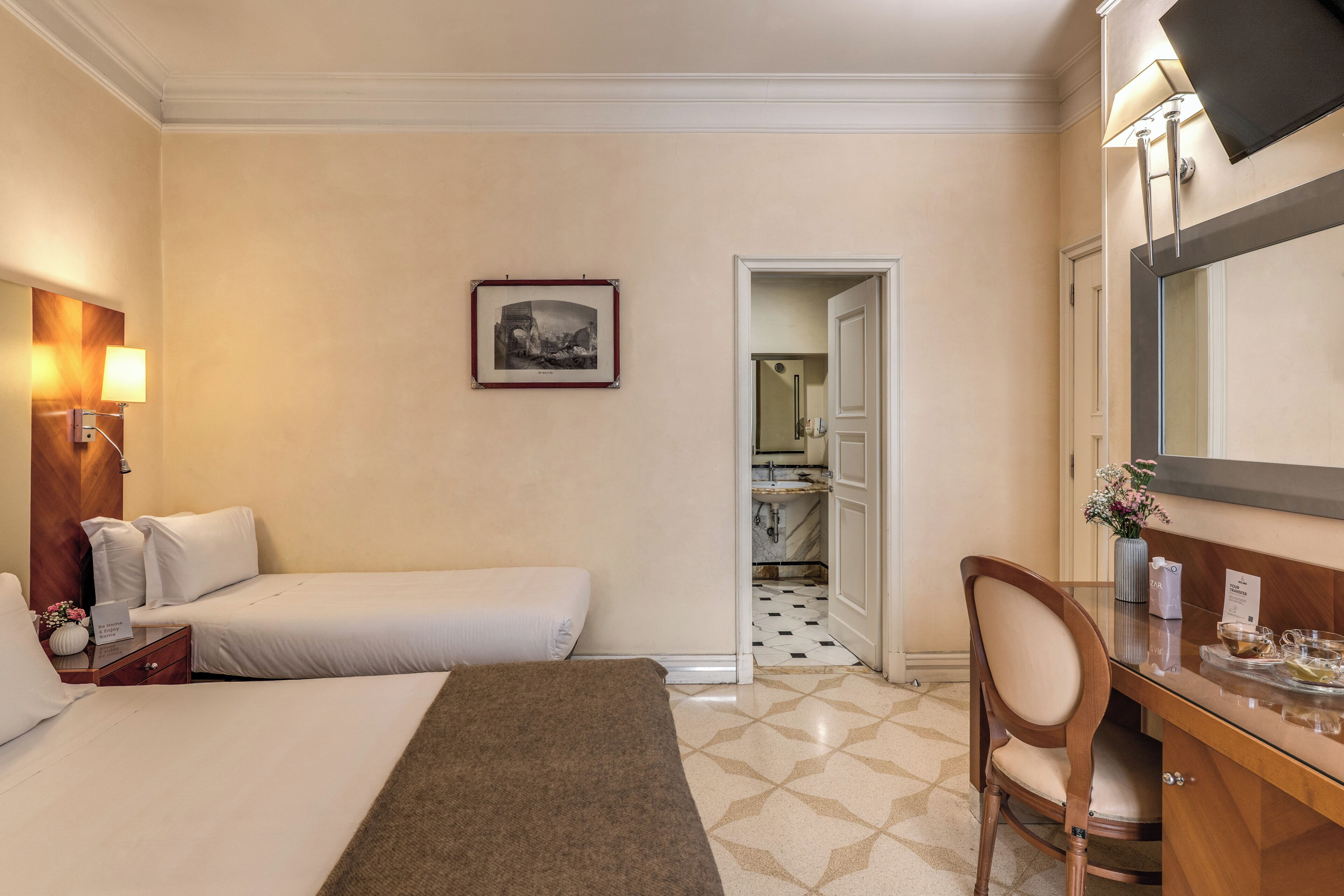 Ein Hotelzimmer mit zwei Betten, einem Schreibtisch mit Stuhl, einem Spiegel, einem gerahmten Bild an der Wand und einem durchgegangenen Tür erkennbaren Badezimmer.