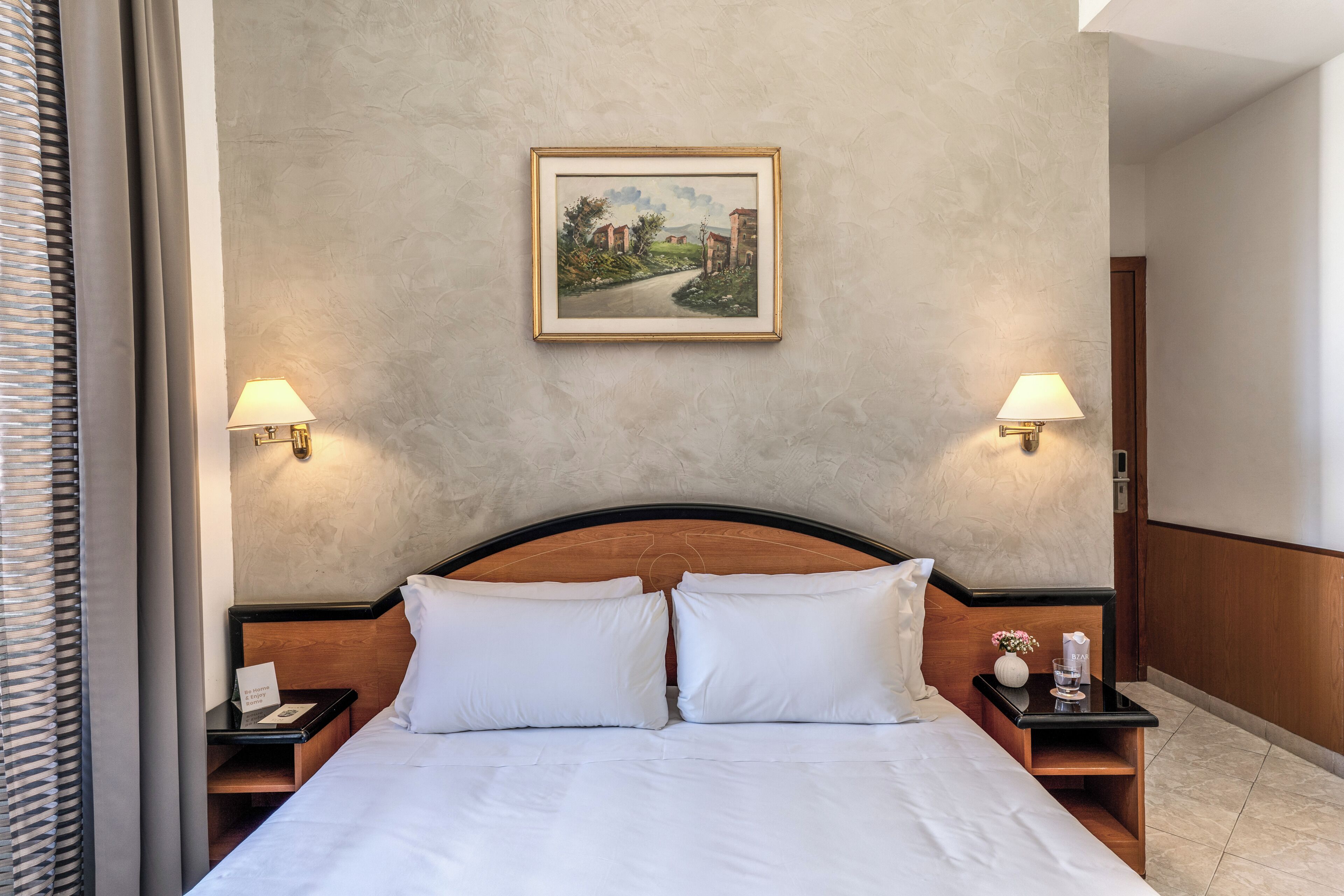 A hotel room with a bed, two bedside tables, a nightstand, a framed picture on the wall, and two wall-mounted lamps.