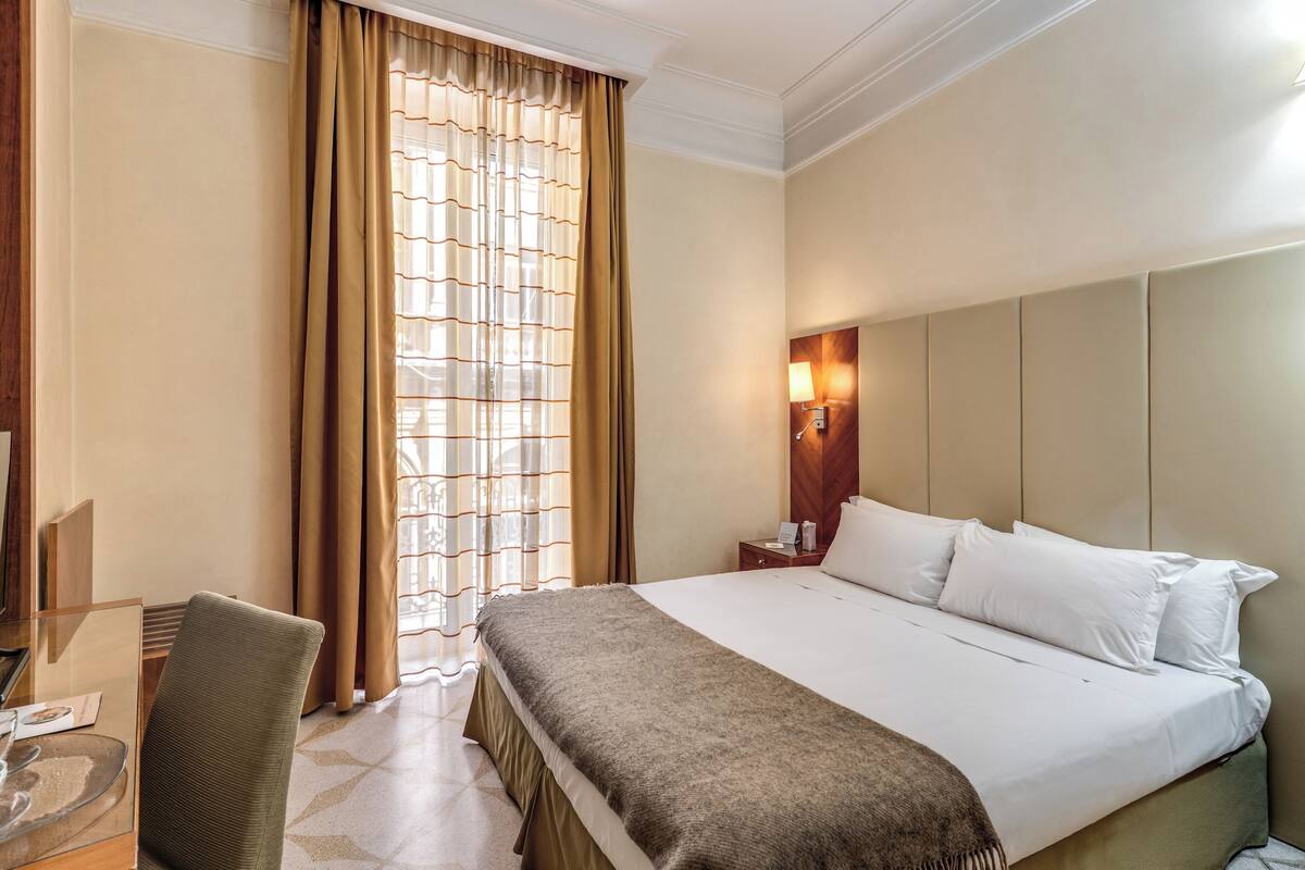 A hotel room with a large bed, a desk with a chair, a lamp, and a window with curtains.