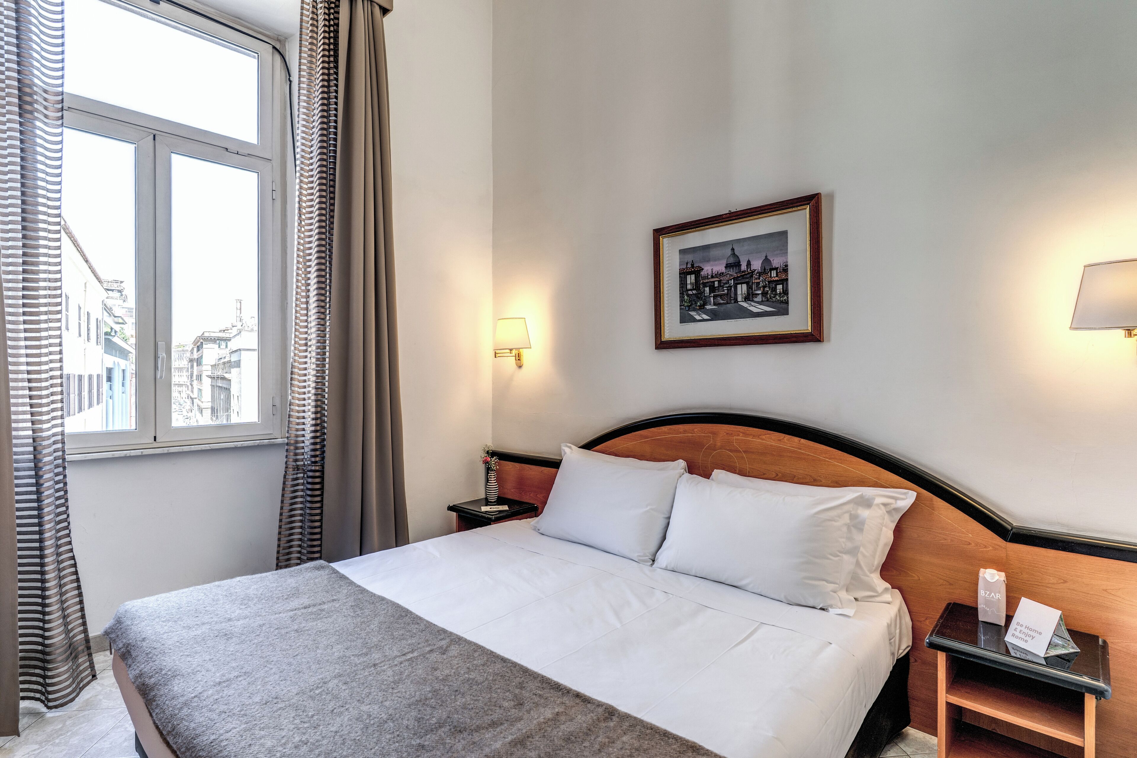Una habitación de hotel con una cama, una mesita de noche, un cuadro enmarcado en la pared y una ventana con cortinas.
