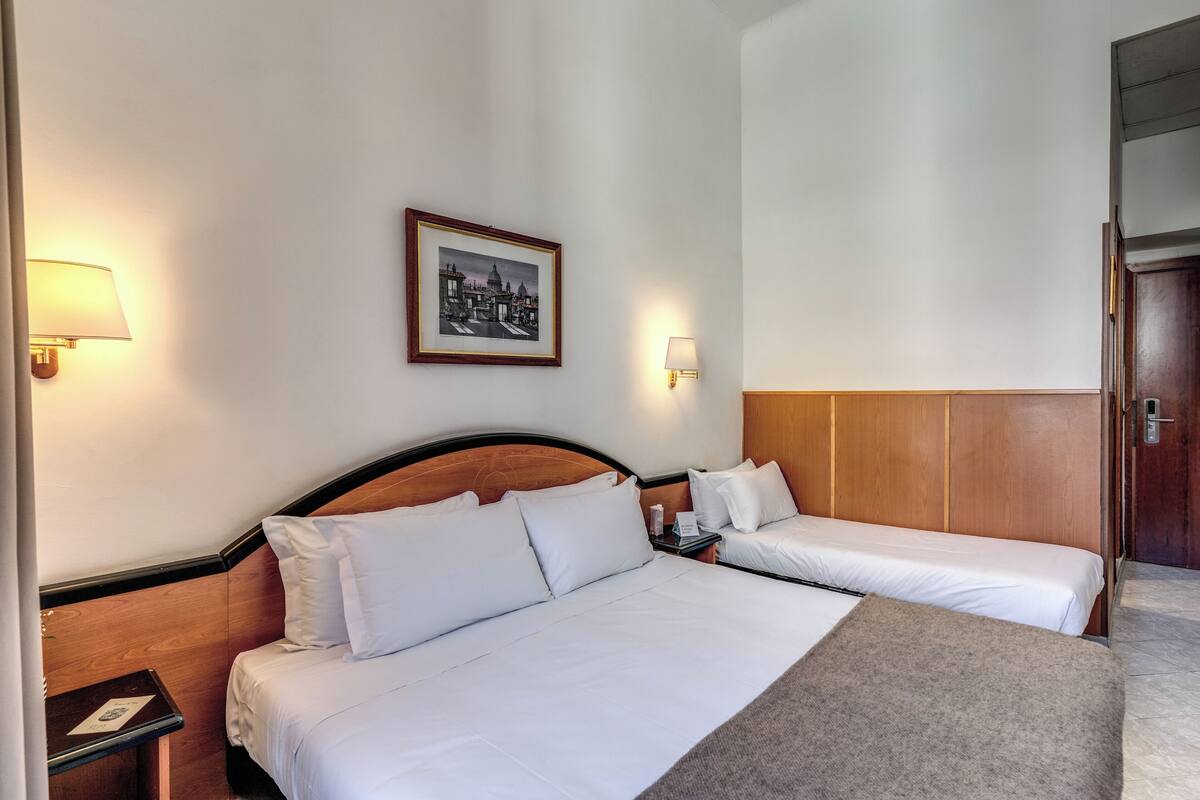 A hotel room with two beds, a nightstand, a lamp, a framed picture on the wall, and a wooden wardrobe.