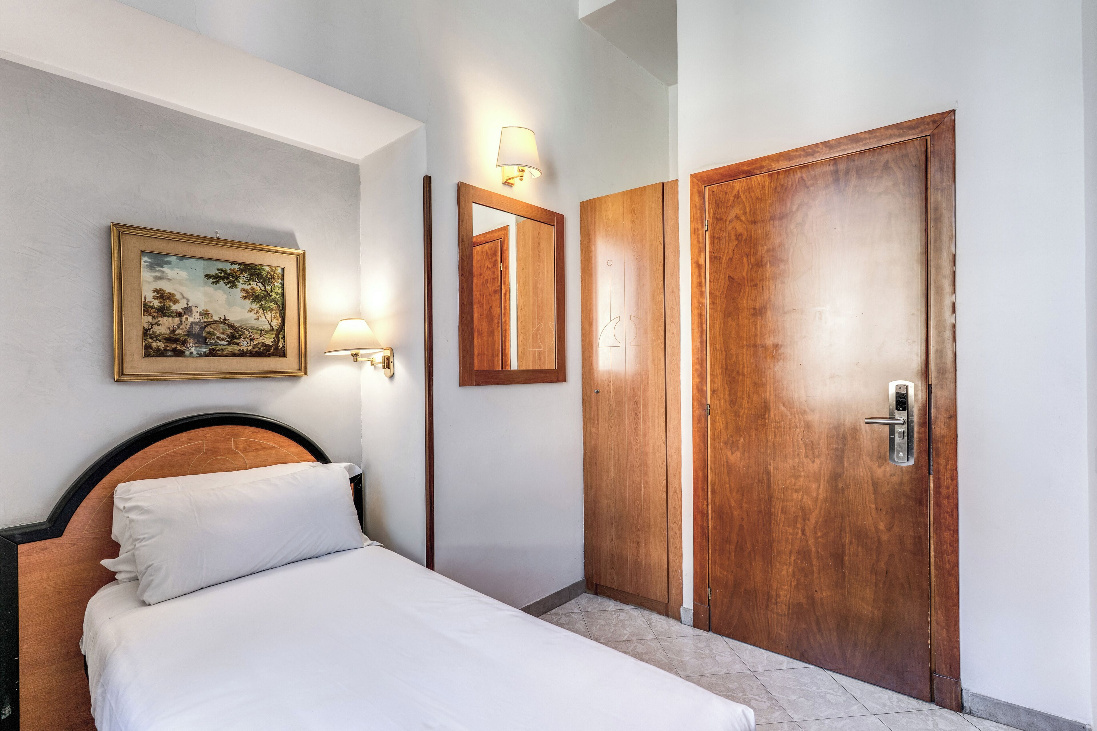 A hotel room with a bed, a wooden wardrobe, a wall-mounted lamp, and a framed picture.