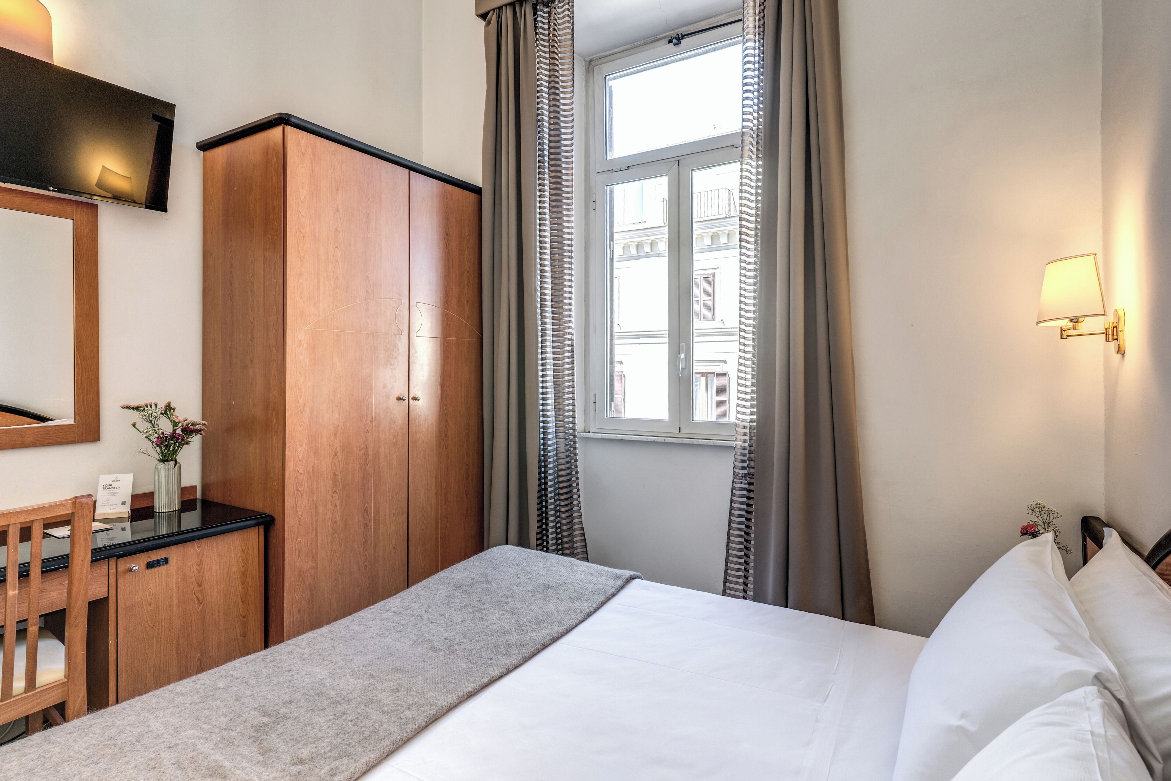 Une chambre d’hôtel comprenant un lit, un bureau, une armoire, une fenêtre avec des rideaux et une lampe fixée au mur.