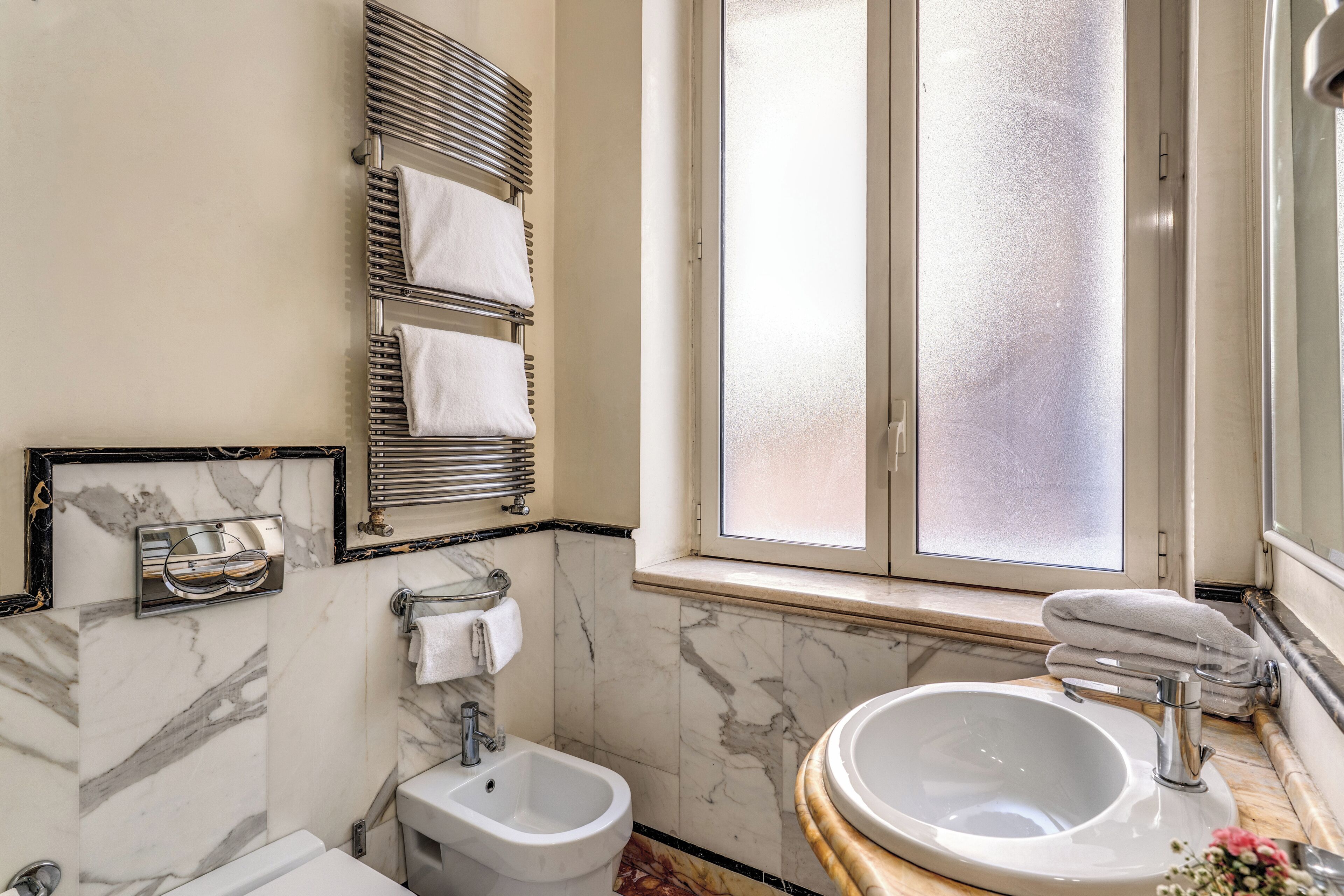 A bathroom with a bidet, a sink, a mirror, and a window with frosted glass.