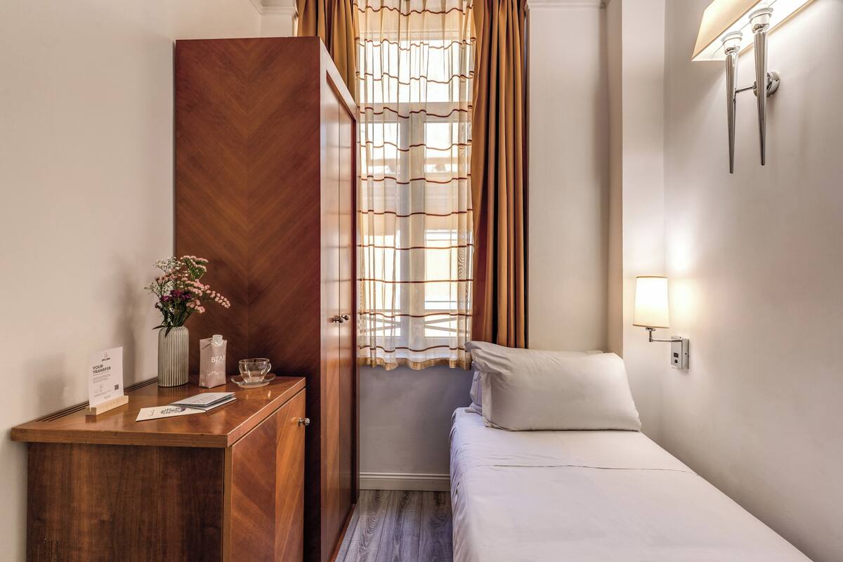 A hotel room with a wooden desk, a bed, a nightstand, a vase with flowers, and a window with curtains.