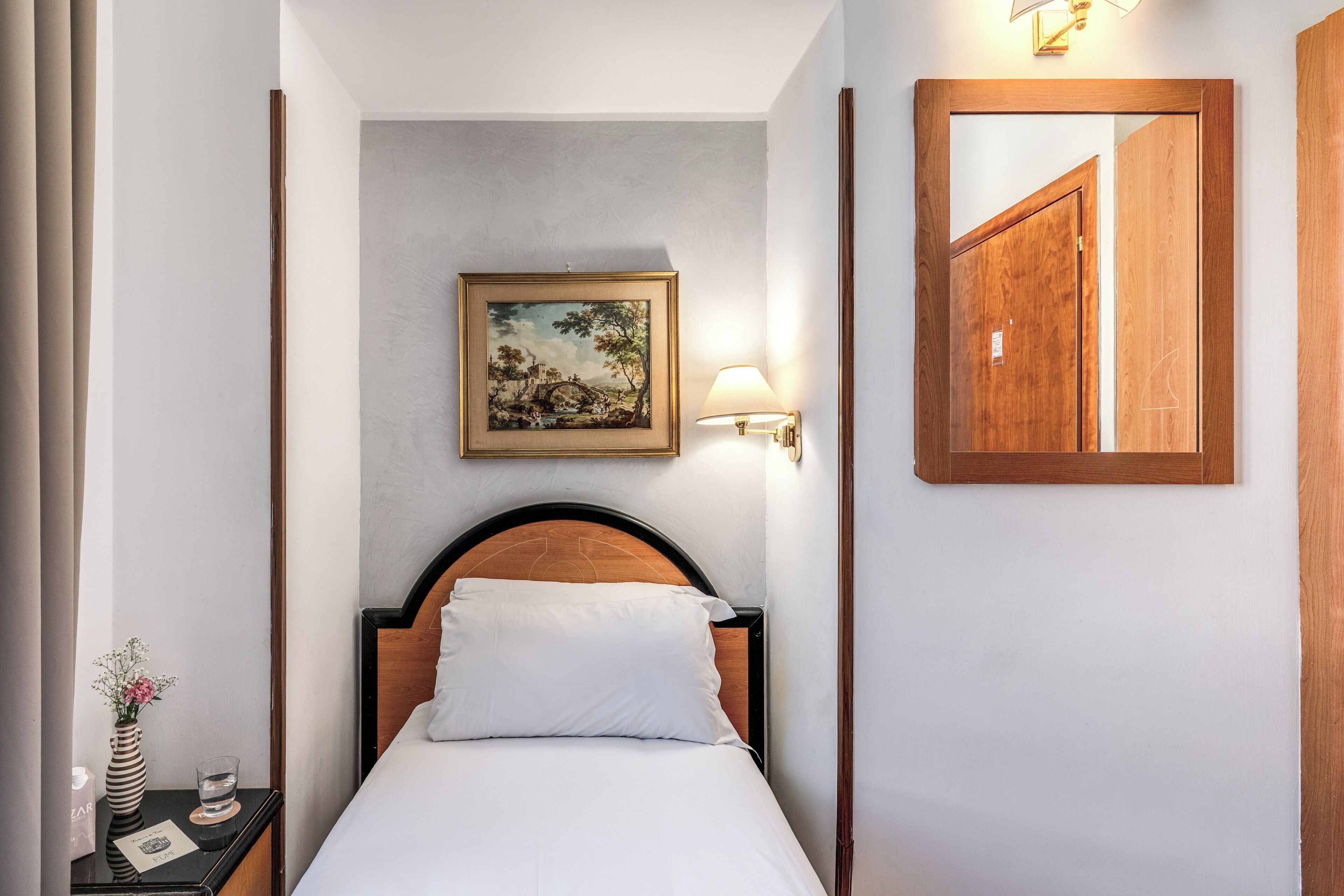 A hotel room with a bed, a nightstand with a vase and a glass, a framed painting on the wall, and a mirror on the wall.