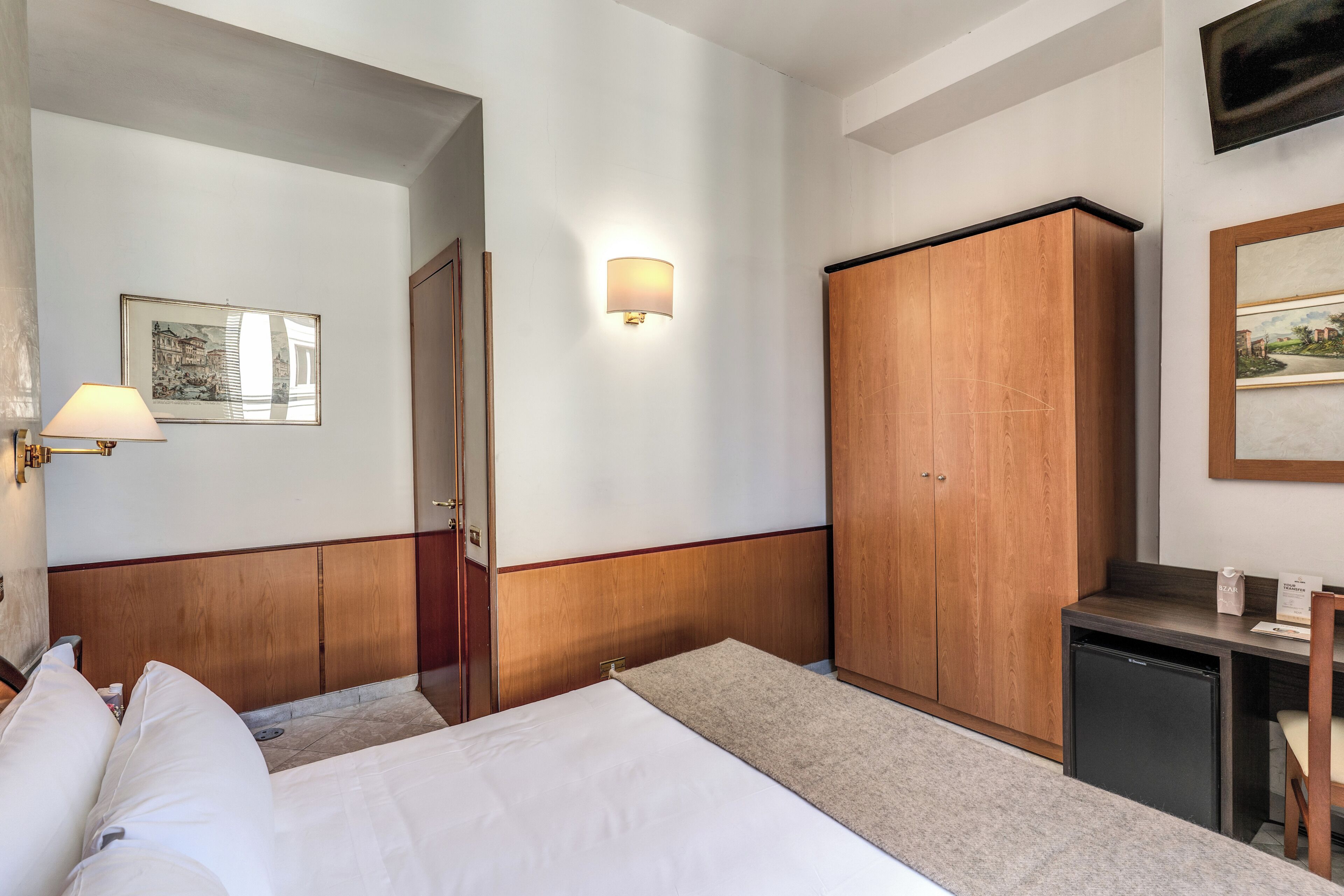 A hotel room with a bed, a desk, a wardrobe, a wall-mounted lamp, and a framed picture.