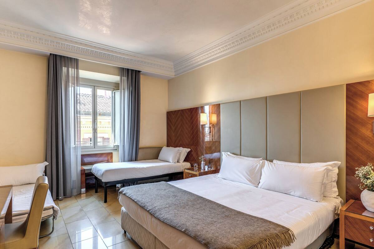 A hotel room with two beds, a desk, a chair, a window with curtains, and a wooden headboard.