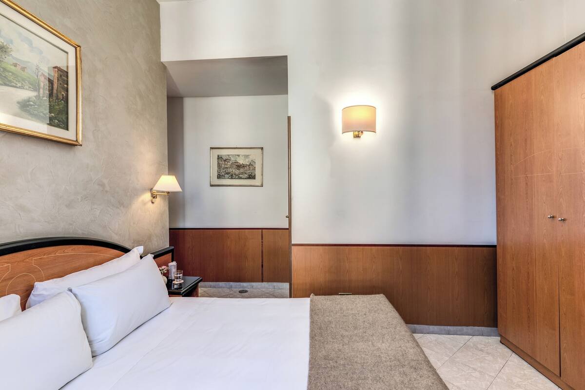 A hotel room with a bed, a nightstand, a lamp, a wall-mounted light, a wardrobe, and a framed picture on the wall.