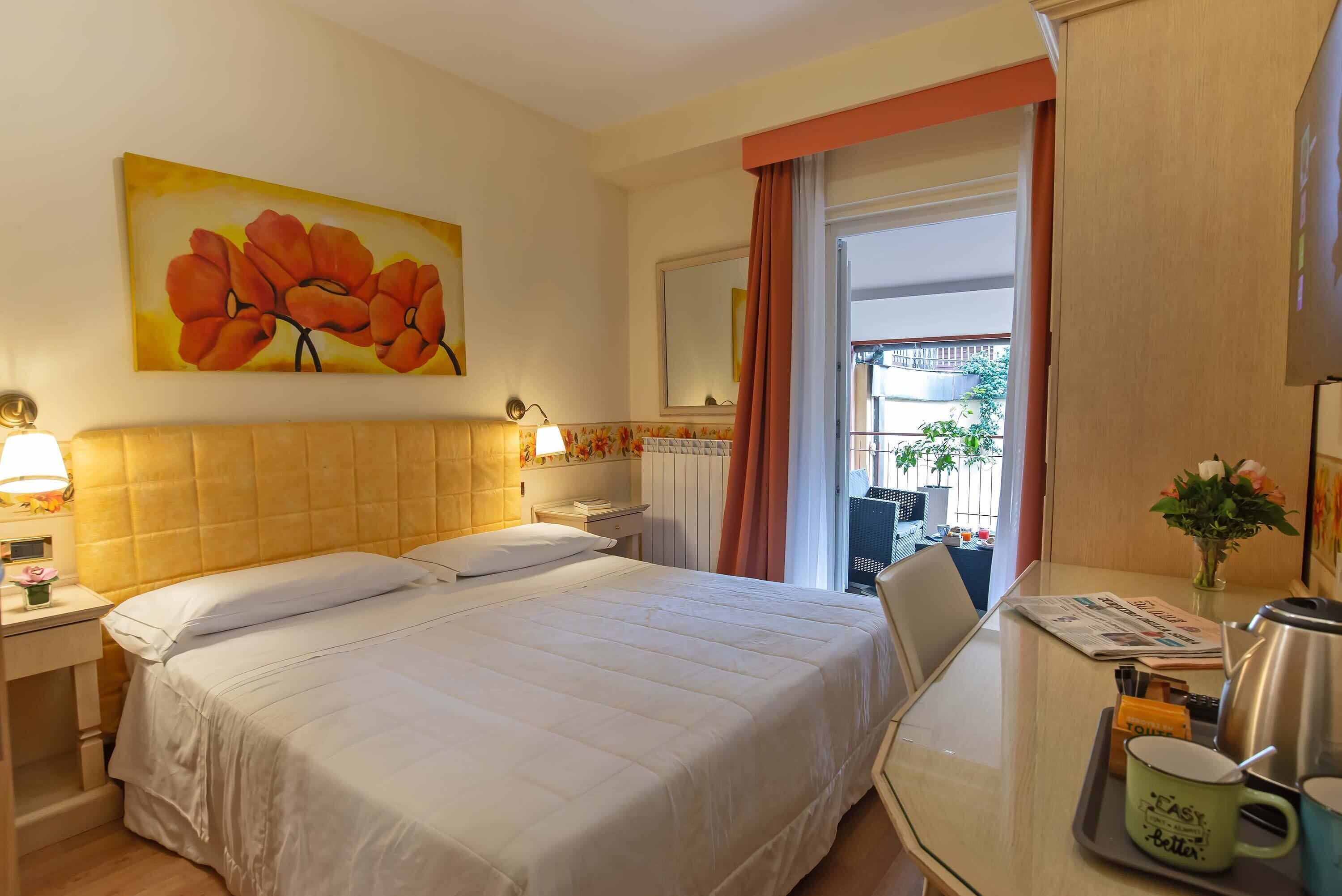 A hotel room with a bed, bedside tables, a desk with a chair, a window with curtains, and a painting on the wall.