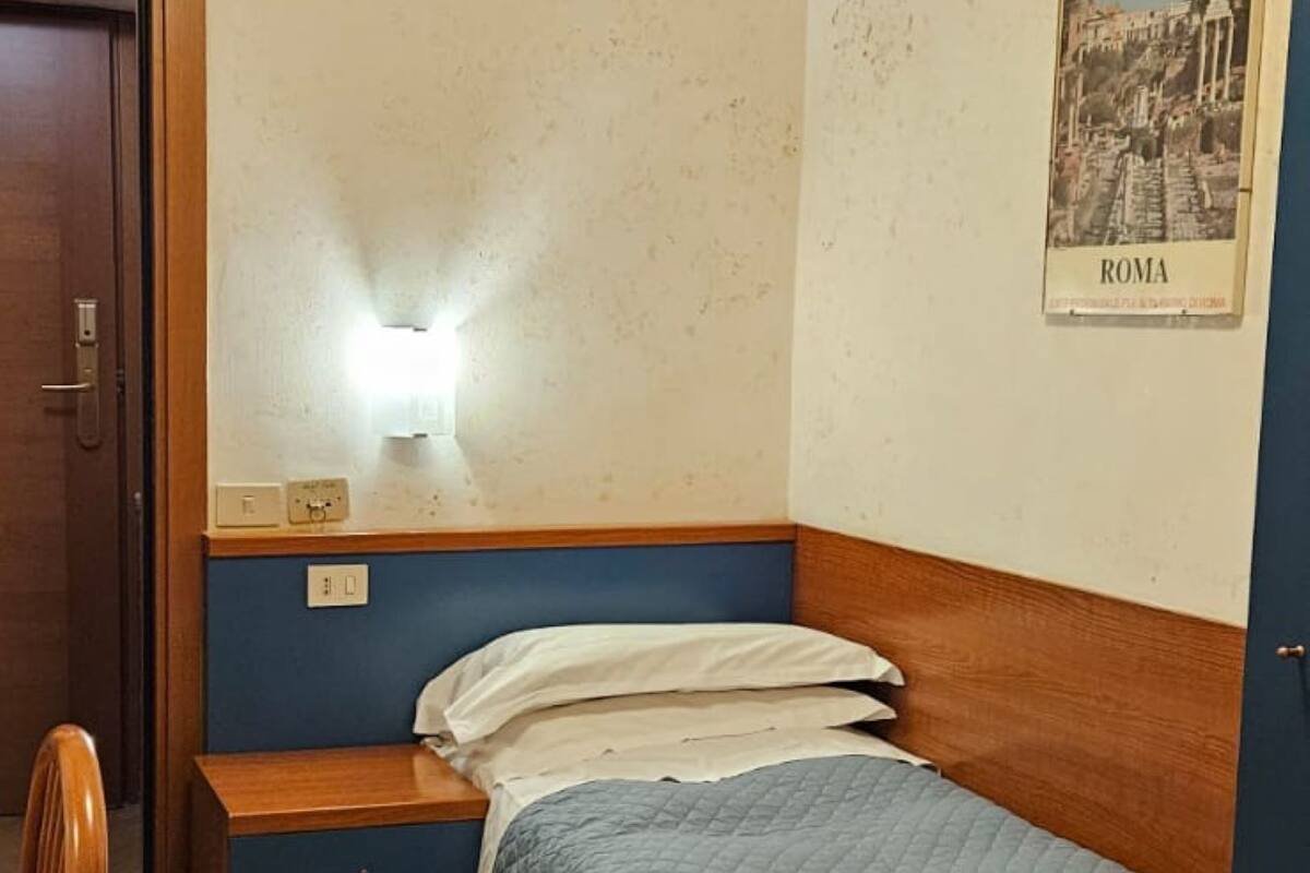 Un dormitori petit i ben organitzat amb un llit individual, una tauleta de nit, una cadira i una làmpada penjada a la paret.