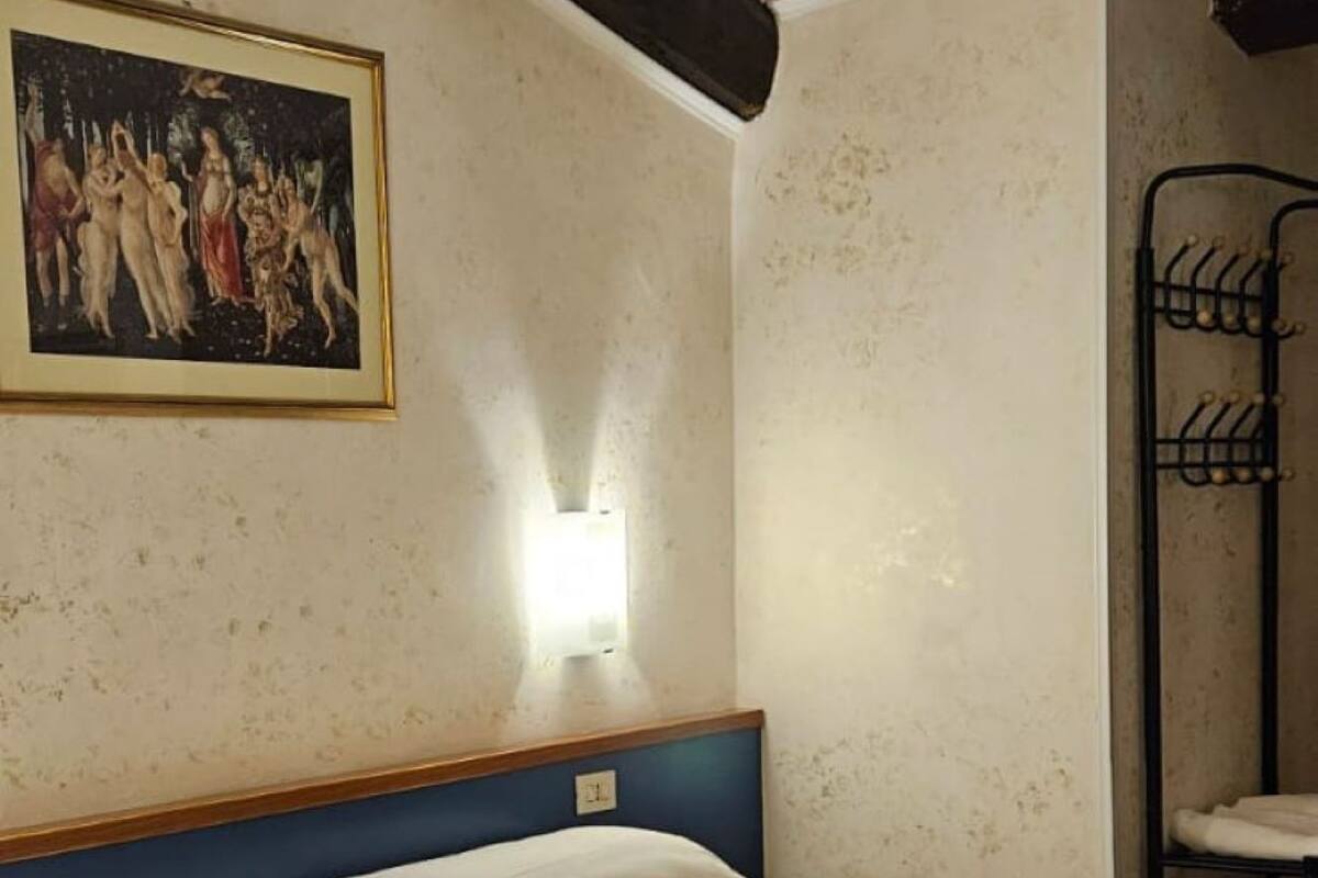 Un dormitori amb un llit, una làmpada penjada a la paret, un quadre i una prestatgeria de metall.