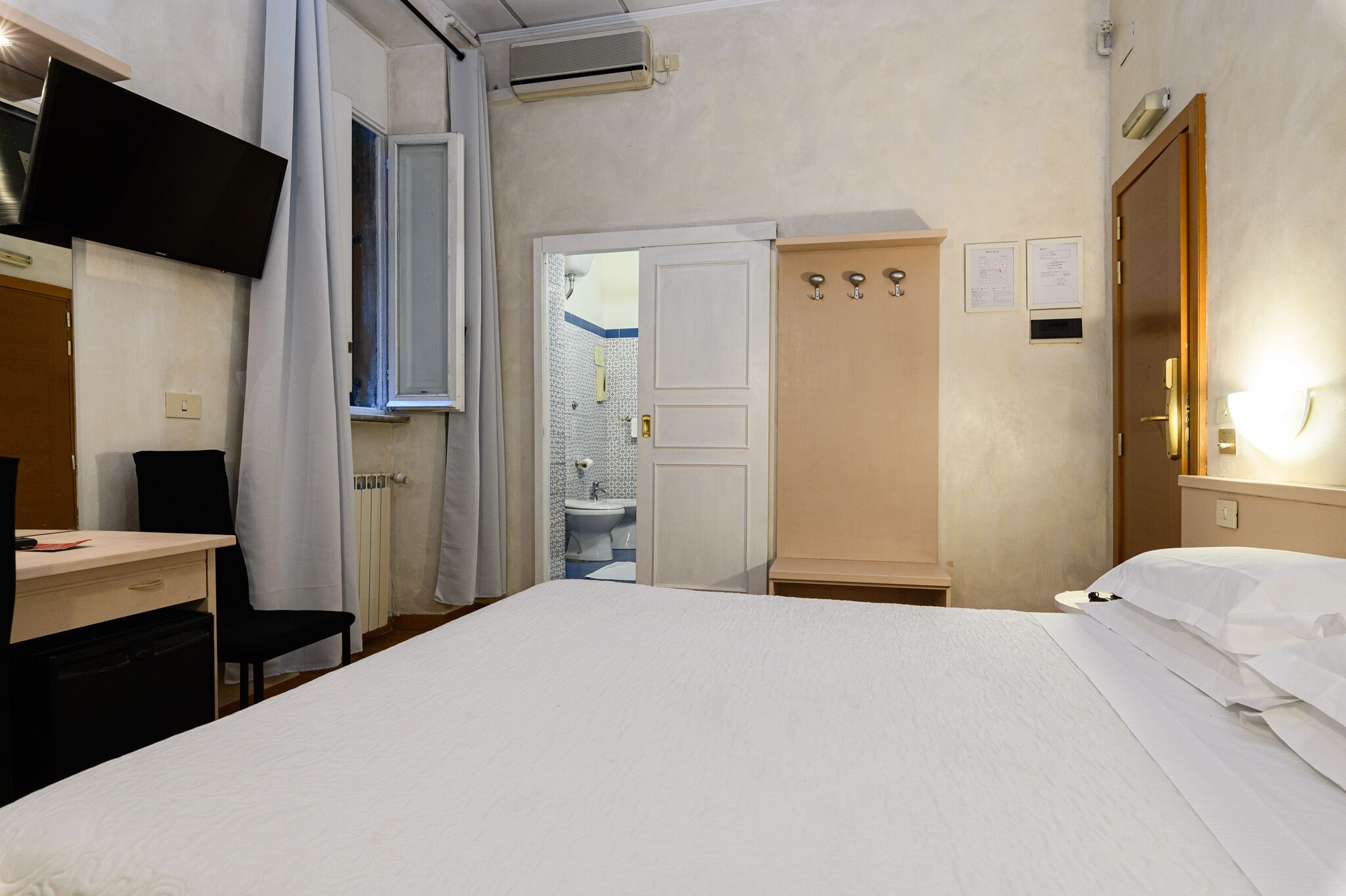 A hotel room with a bed, a desk, a chair, a wardrobe, and a bathroom visible through an open door.