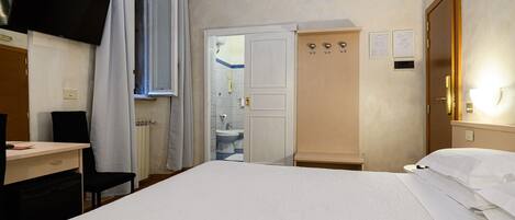 A hotel room with a bed, a desk, a chair, a wardrobe, and a bathroom visible through an open door.