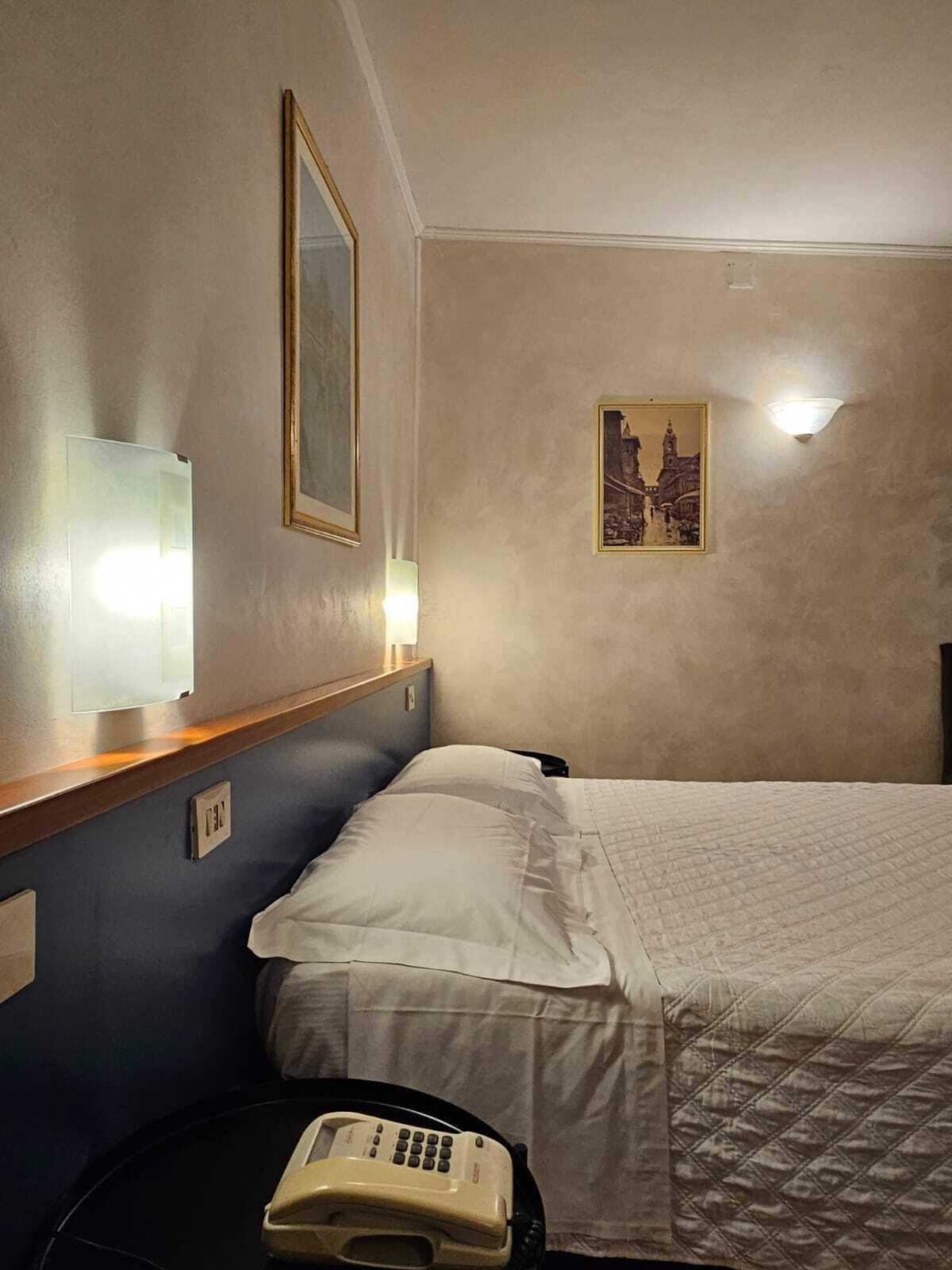 Habitación de hotel con cama, mesita de noche, teléfono, luz empotrada en la pared y un cuadro enmarcado.
