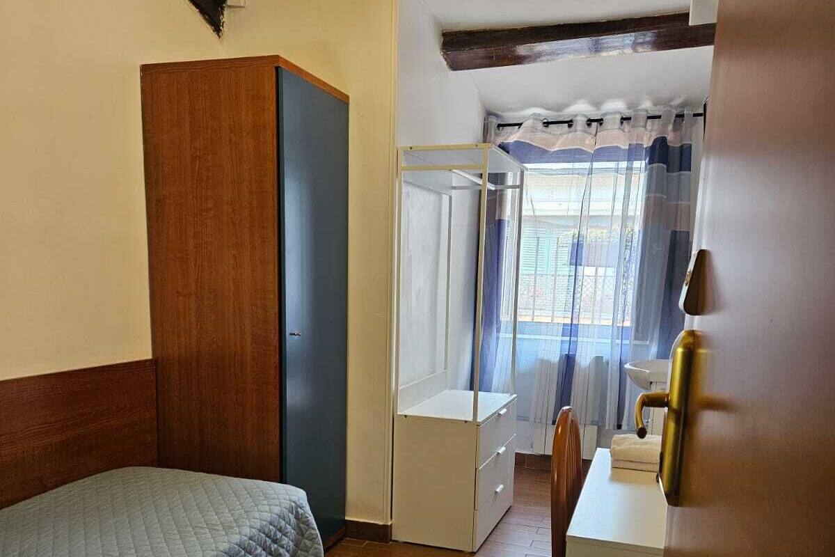 Habitació petita i senzilla amb llit, taula de treball i finestra amb cortines.