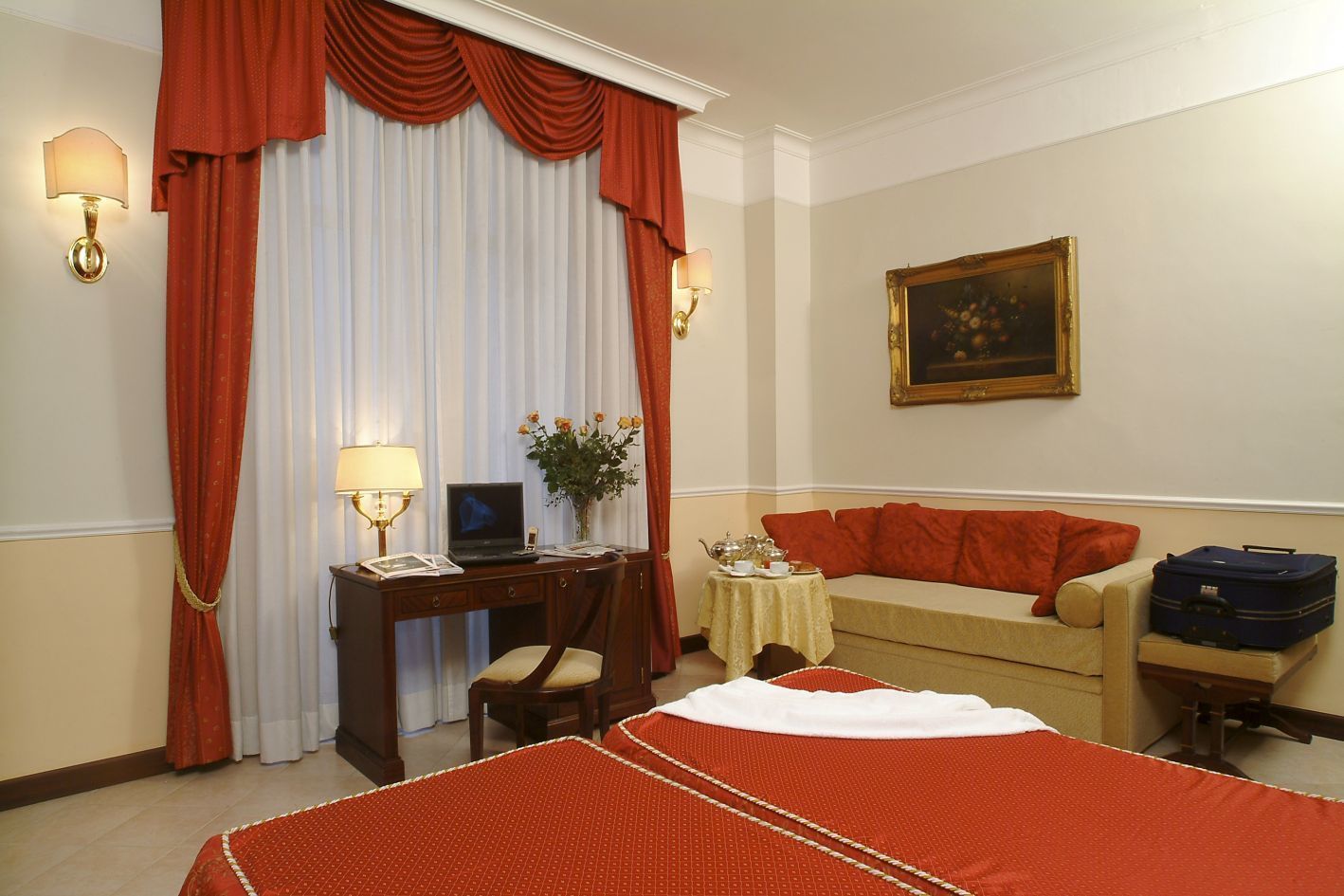 A hotel room with two beds, a desk with a laptop, a small table with a vase of flowers, and a sofa with luggage.