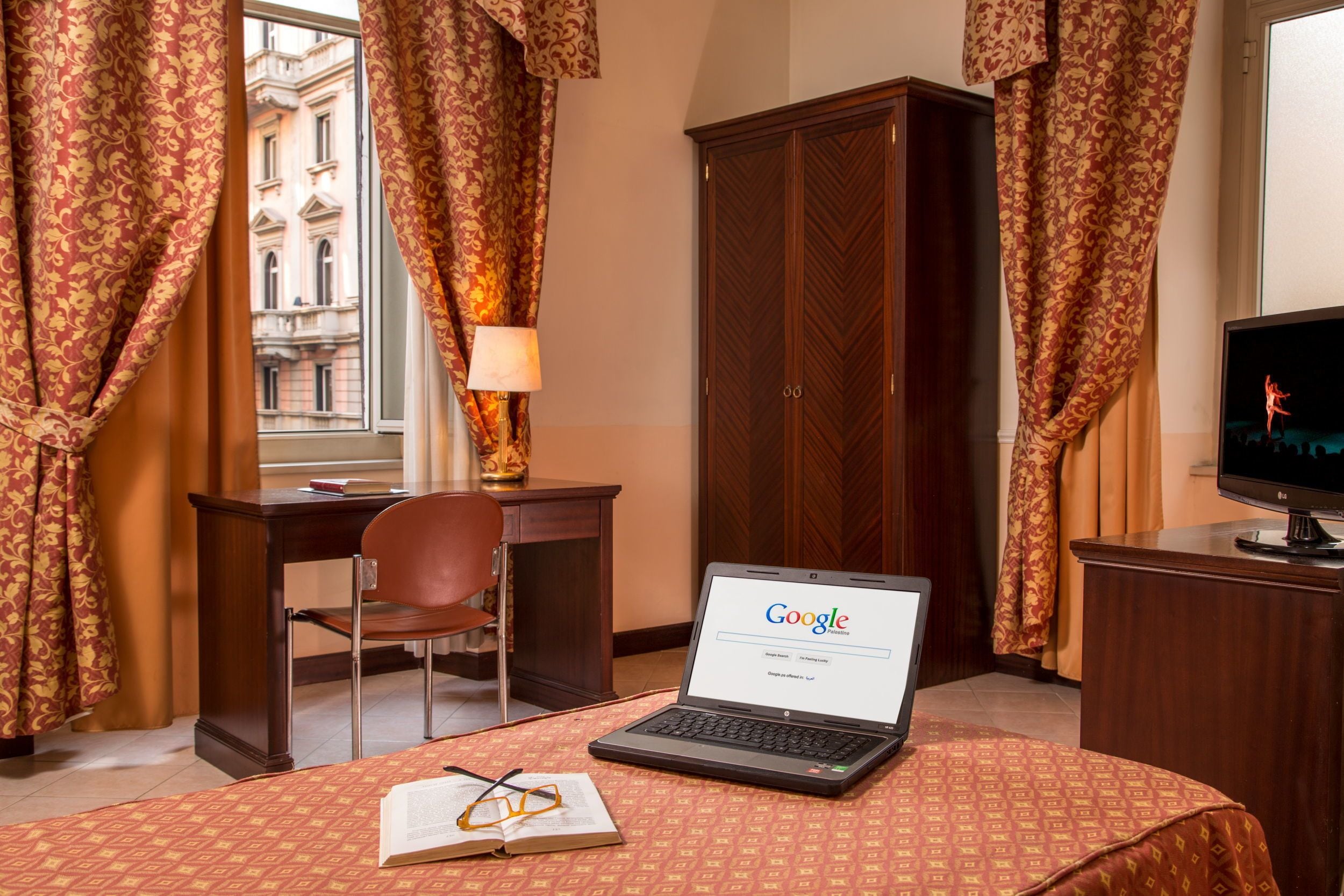 A hotel room with a laptop on a bed, a desk with a chair, a wardrobe, and curtains.