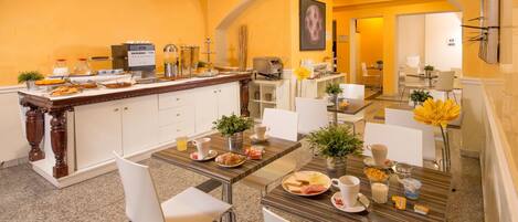Un espace dédié au petit-déjeuner, avec un comptoir en marbre, des chaises blanches et une table à manger dressée avec des assiettes, des tasses et un vase rempli de fleurs.