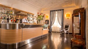 Un espace bar avec un comptoir en bois poli, un barman et une étagère où sont exposées différentes bouteilles. On y trouve également un vase décoratif avec des fleurs et un fauteuil classique.
