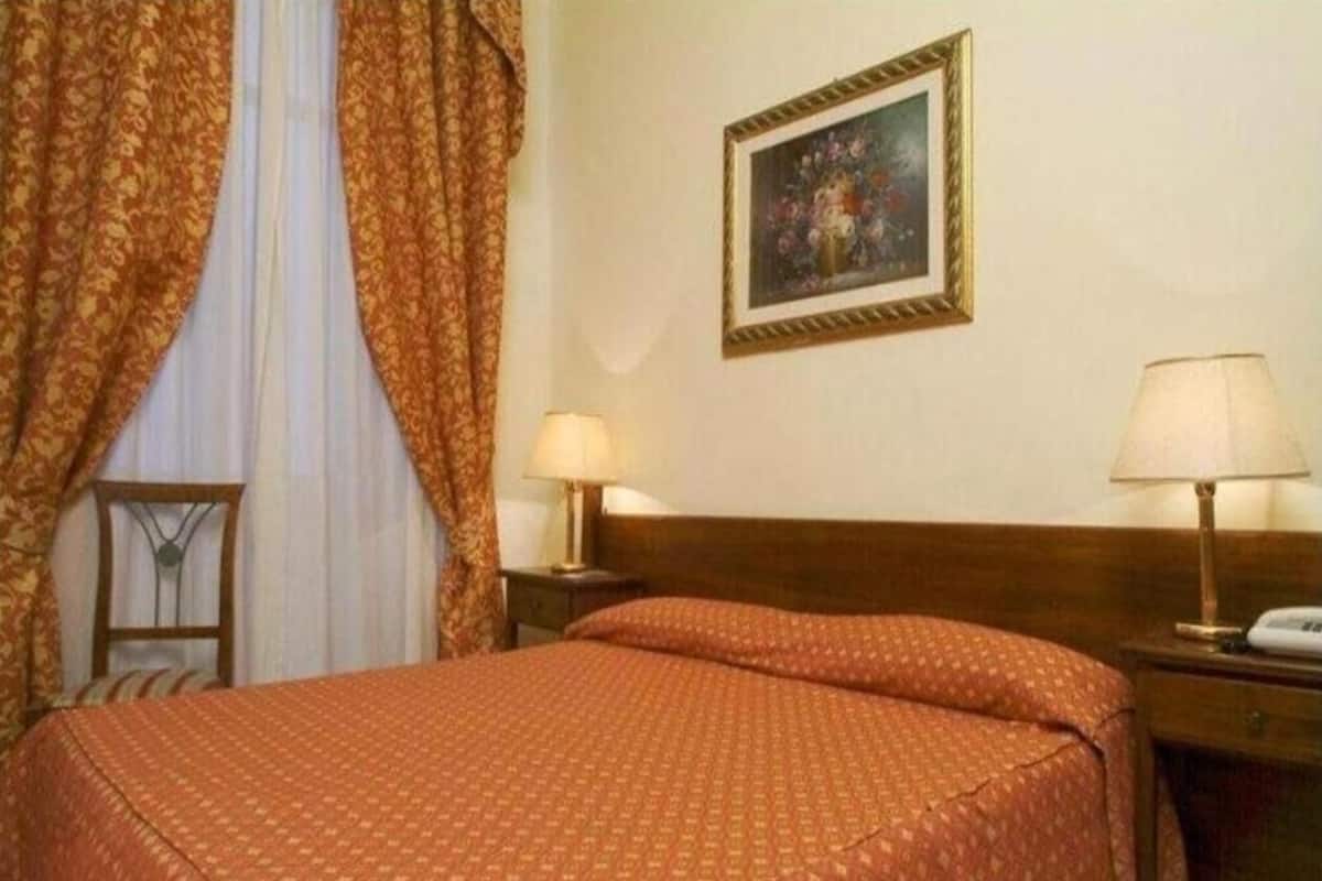 A hotel room with a bed, two bedside lamps, a nightstand, a wooden headboard, a framed picture on the wall, and a window with curtains.