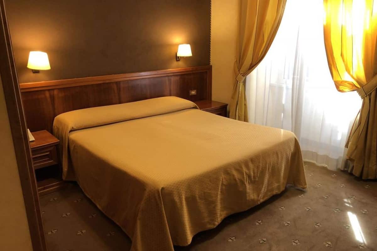 Hotellihuone, jossa on sänky, yöpöydät, puinen pääty ja kaksi seinään kiinnitettyä lamppua.
