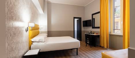 A hotel room with a bed, bedside table, a small desk with a chair, a TV mounted on the wall, and a window with yellow curtains.