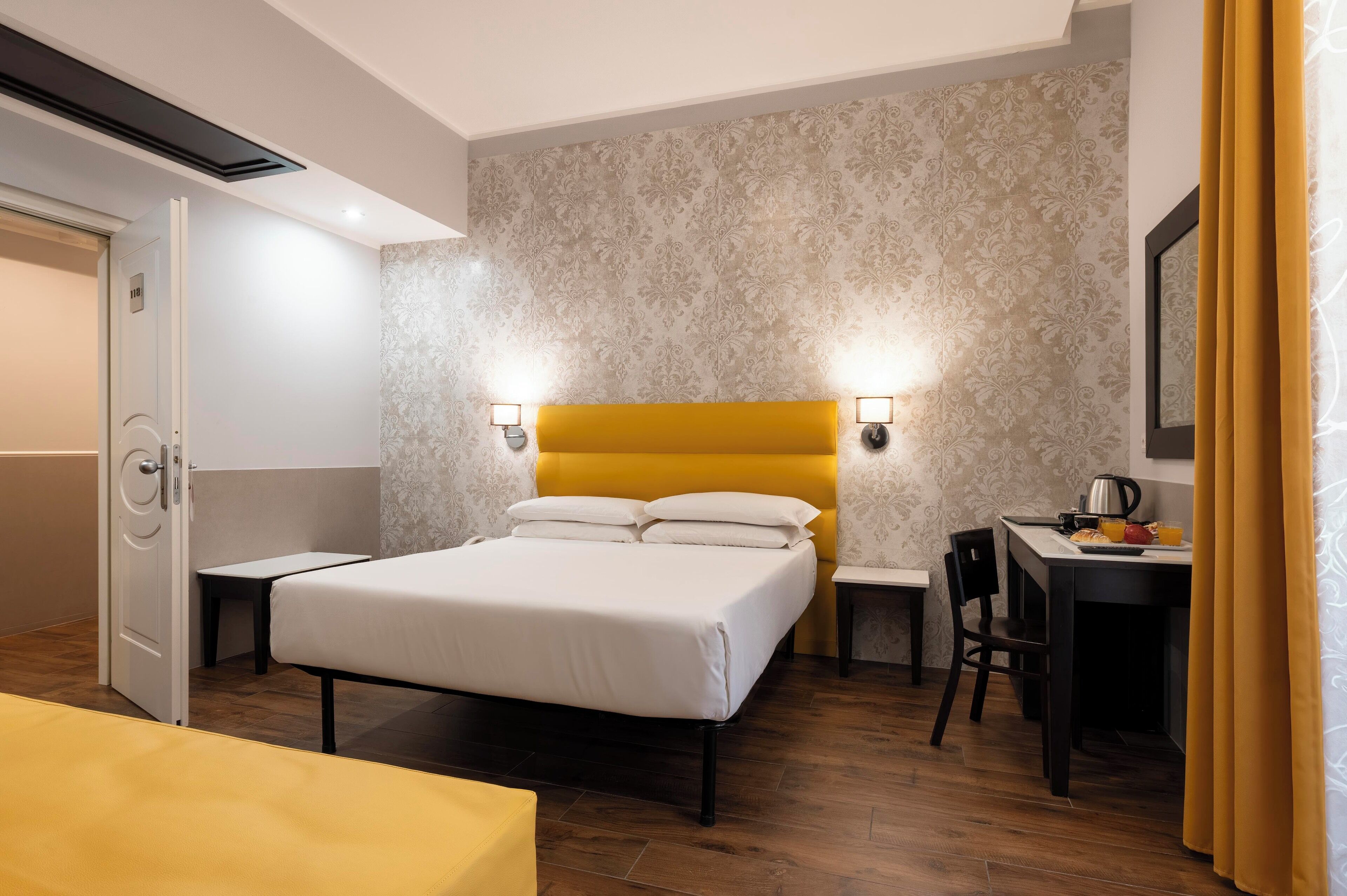 A hotel room with a bed, bedside tables, a desk with a chair, and a small table with a kettle and orange juice.