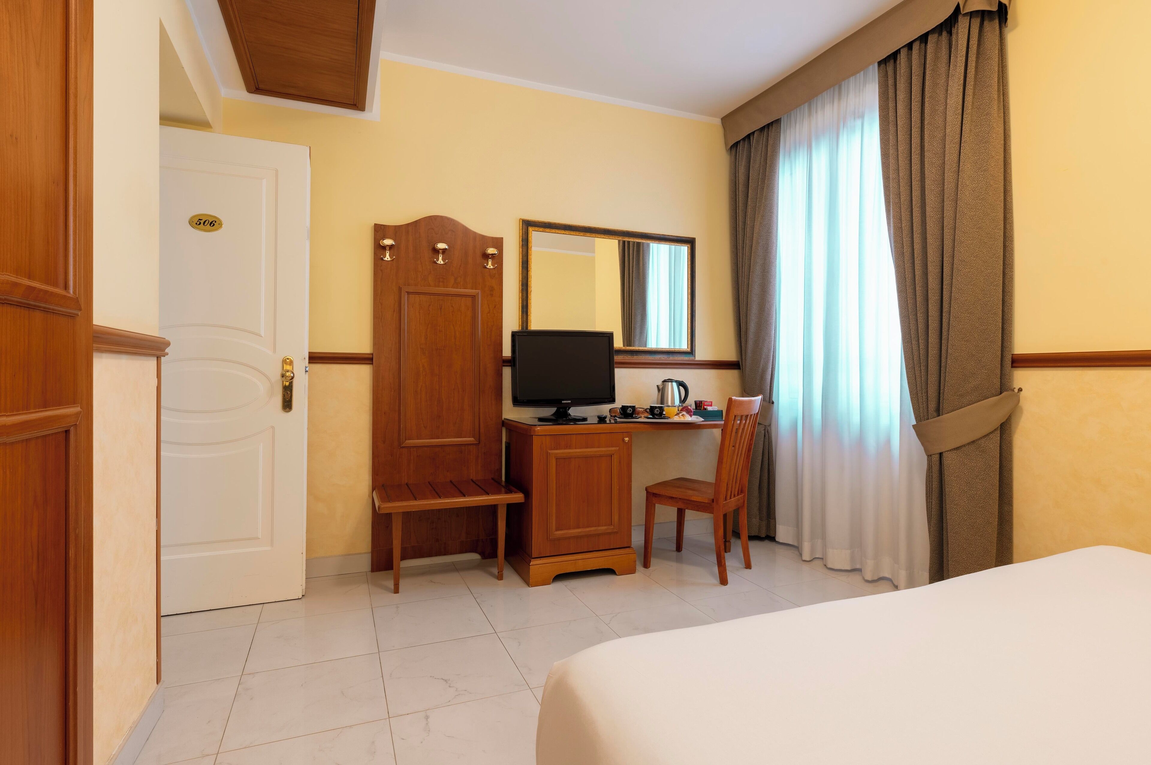 A hotel room with a bed, a desk with a computer, a chair, a wardrobe, and a door with a room number.