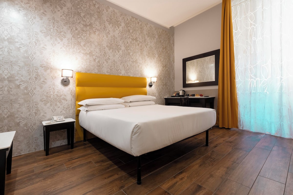 Hotellihuone, jossa on sänky, yöpöydät, yölamppu, peili ja ikkuna, jossa on verhot.