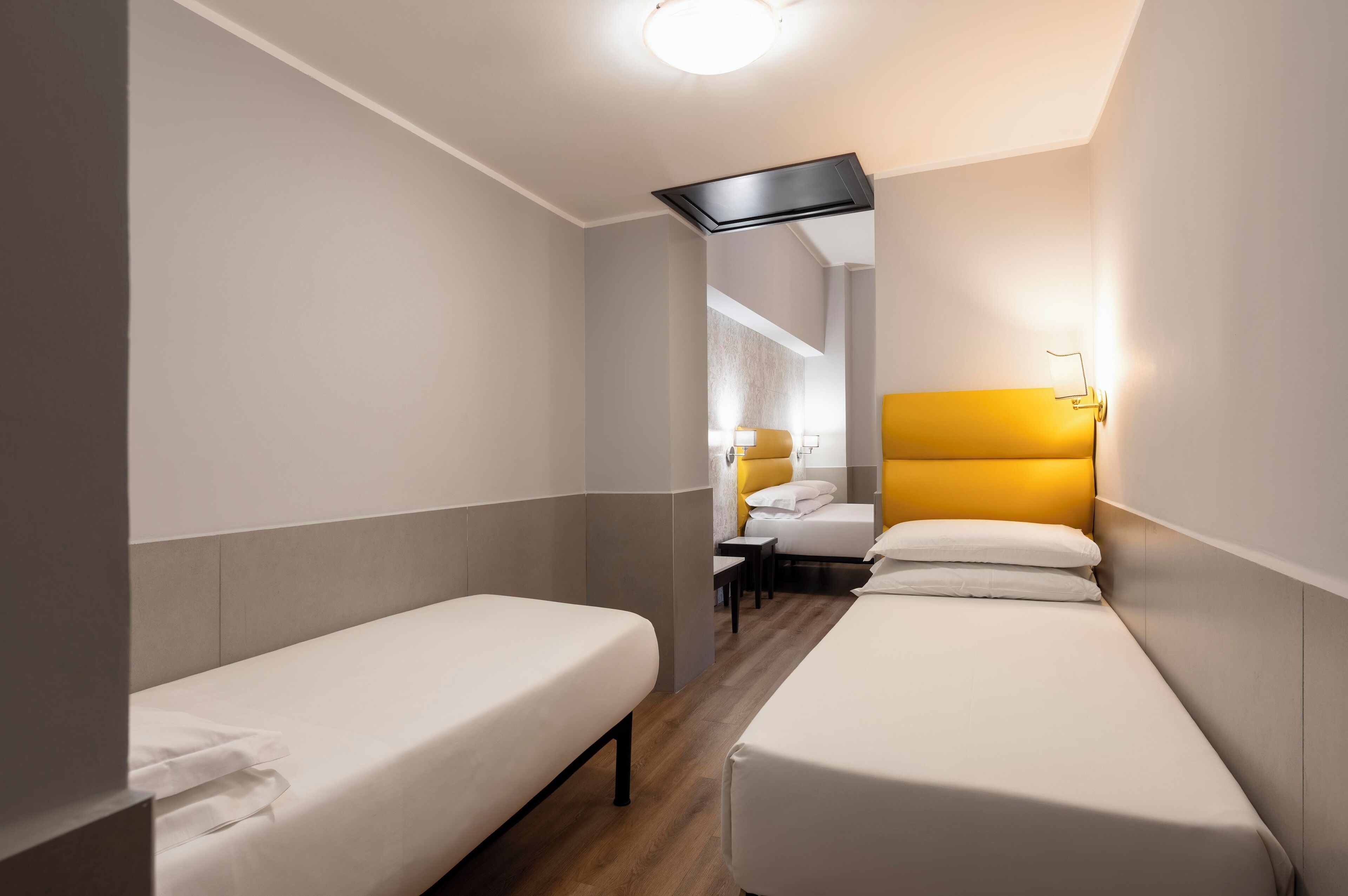 A hotel room with two single beds, a wooden floor, a modern light fixture, and a view of another room with a bed and a nightstand.