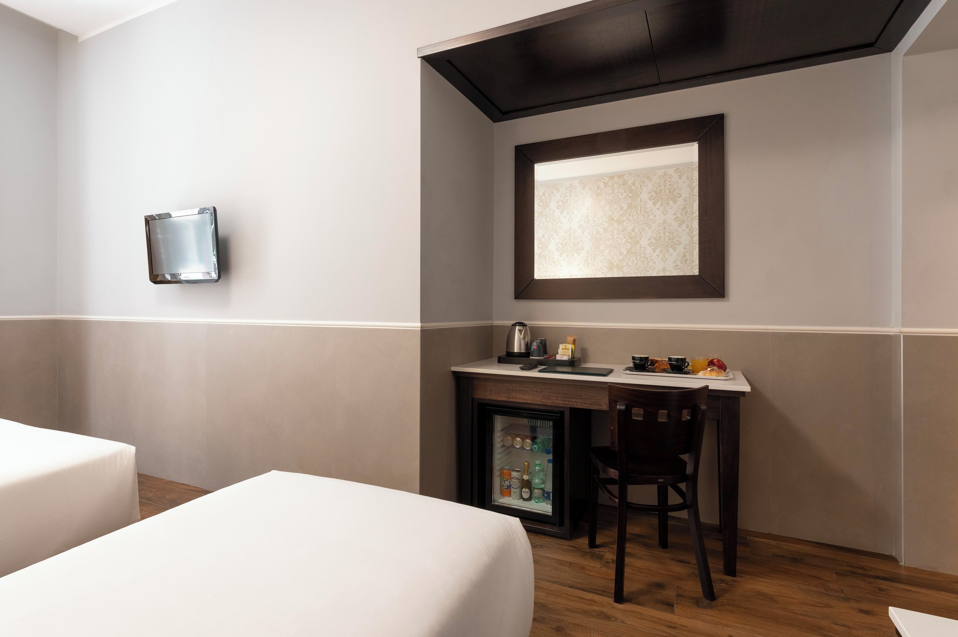 A hotel room with a bed, a desk with a refrigerator, a chair, and a wall-mounted light fixture.