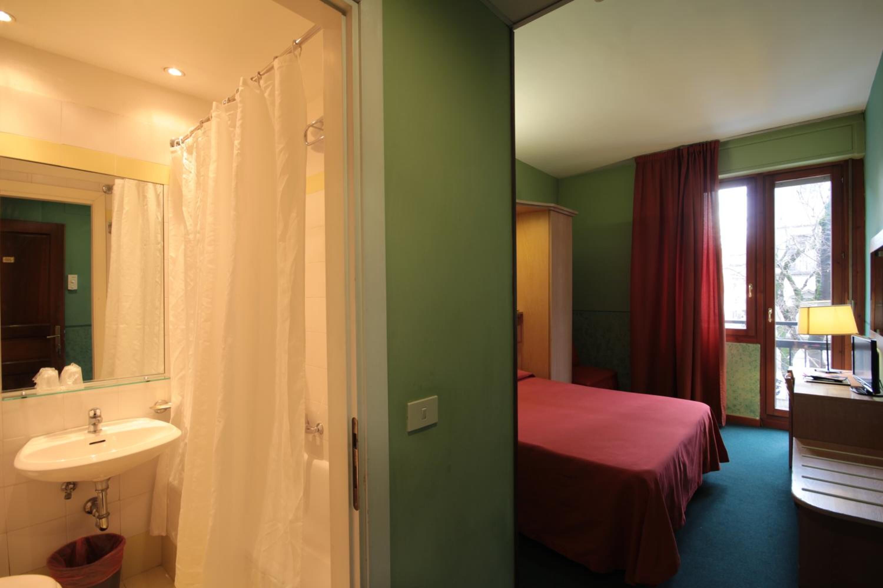 A hotel room with a bathroom, a bed, a desk with a lamp, and a window with curtains.