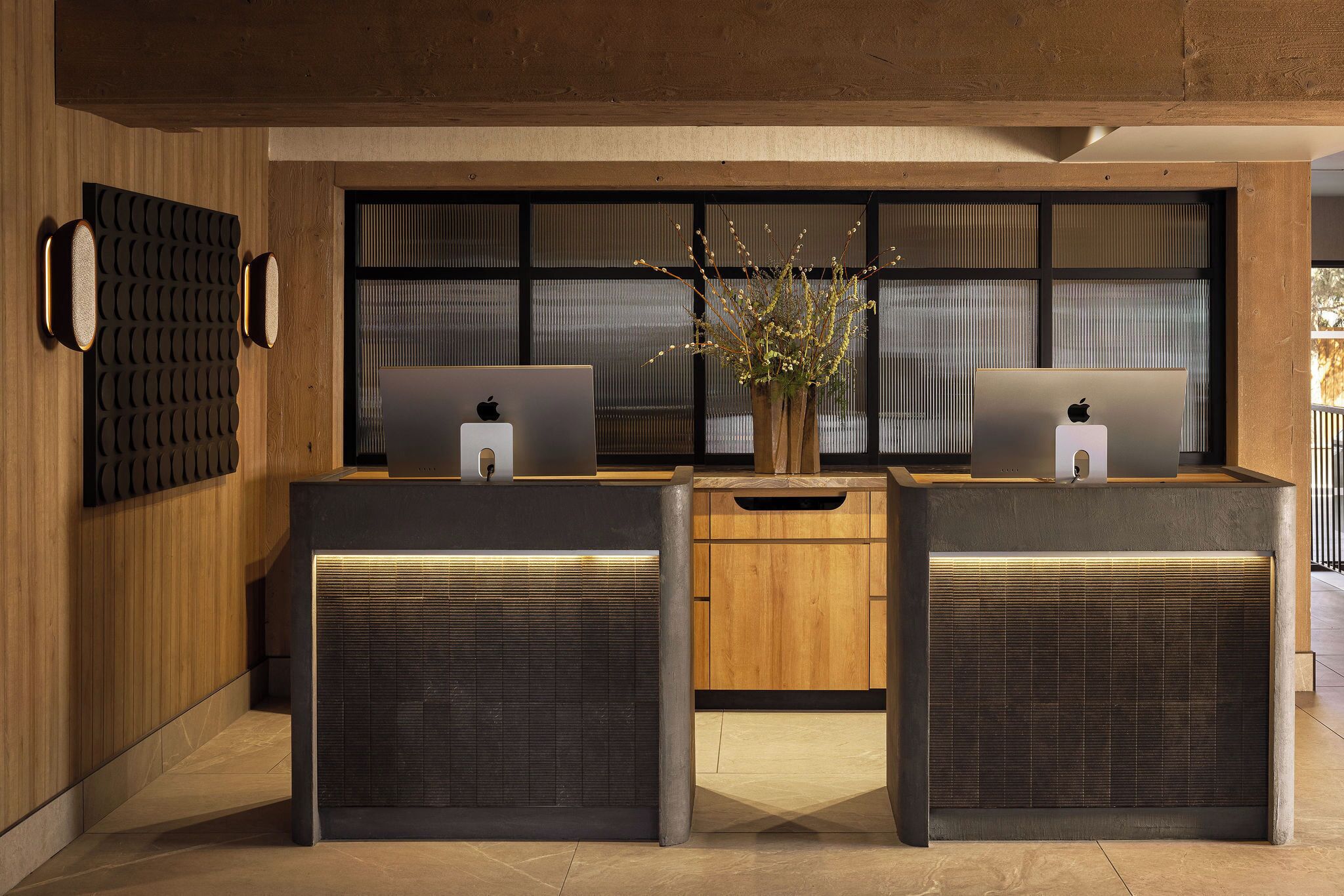 A reception desk with two Apple computers, a vase with flowers, and a wooden cabinet.