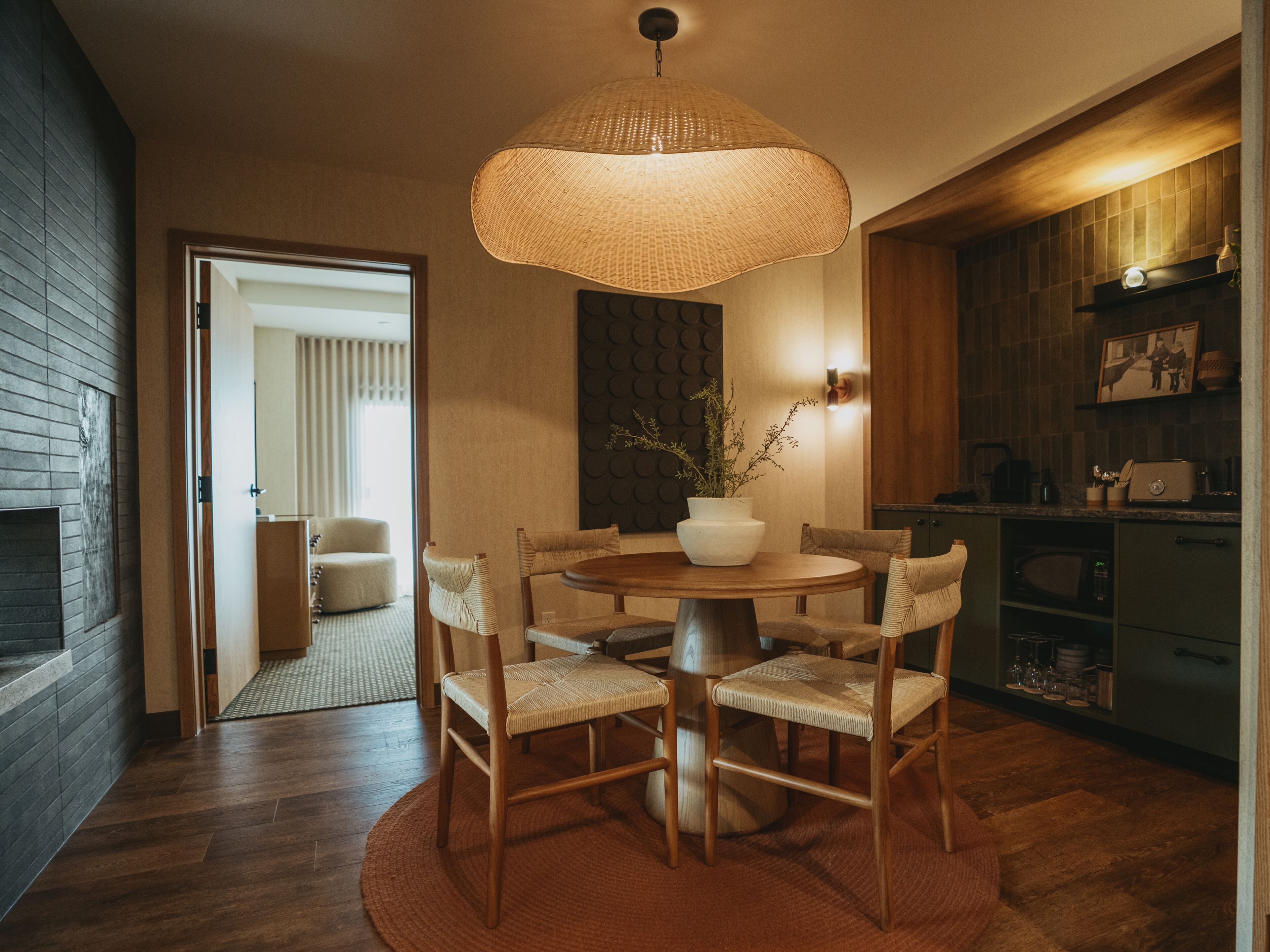 A dining area with a round table, six chairs, a woven pendant light, a vase with plants, and a kitchen in the background.