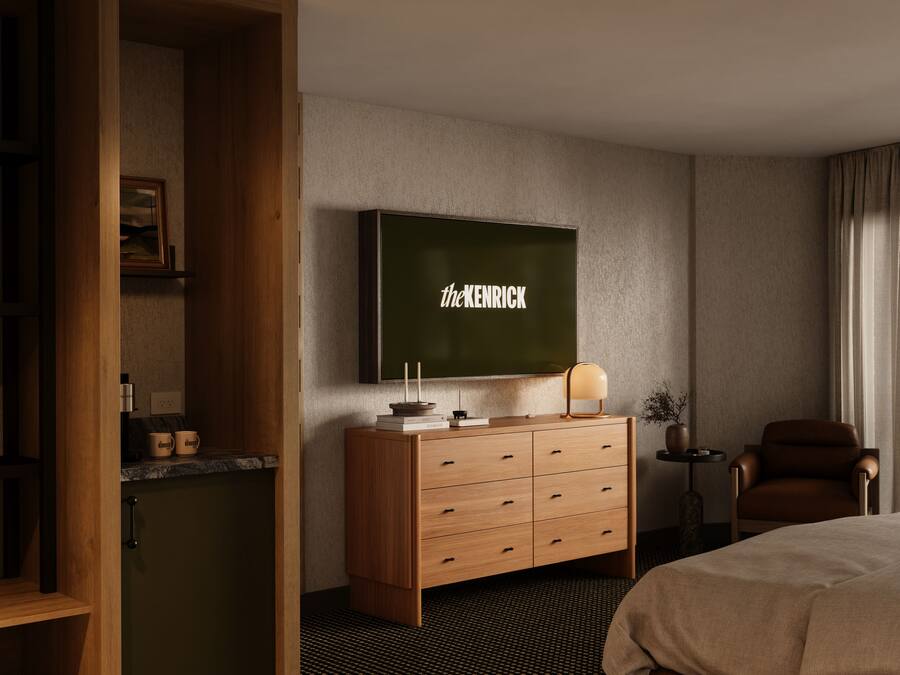 Een hotelkamer met een grote flatscreen televisie aan de muur, een houten dressoir met lades, een klein bijzettafeltje met een lamp, een bruine leren fauteuil en een bed met een beige deken.